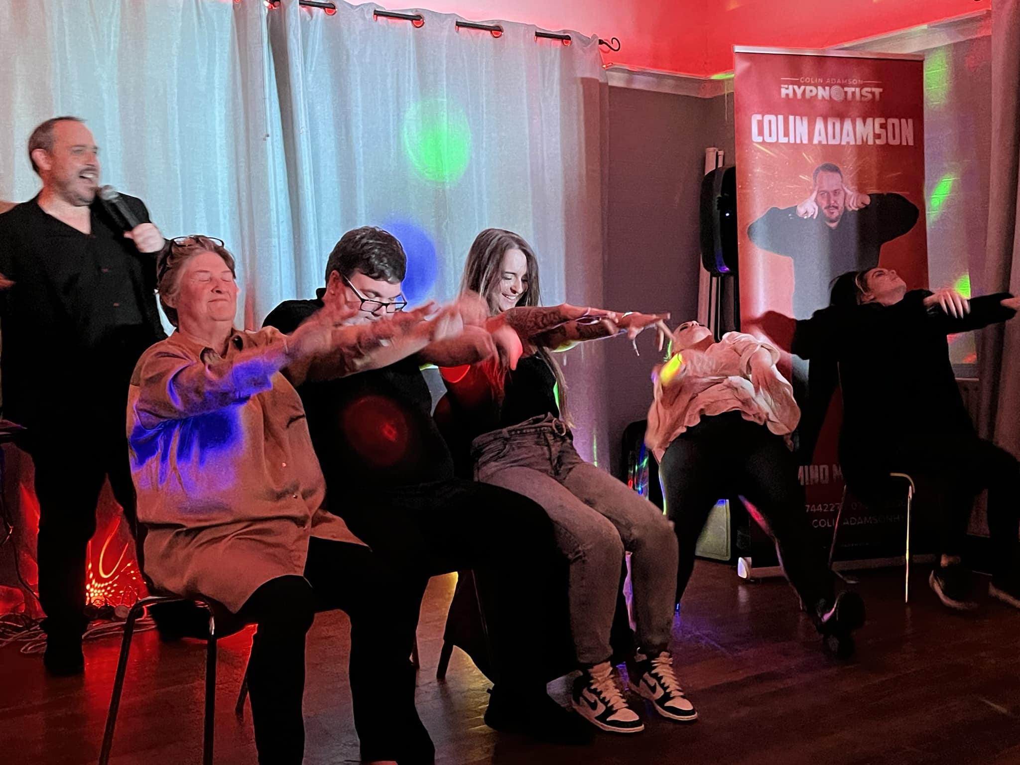 A lively hypnotism show features participants in various relaxed poses under vibrant colored lights, creating an engaging and entertaining atmosphere.