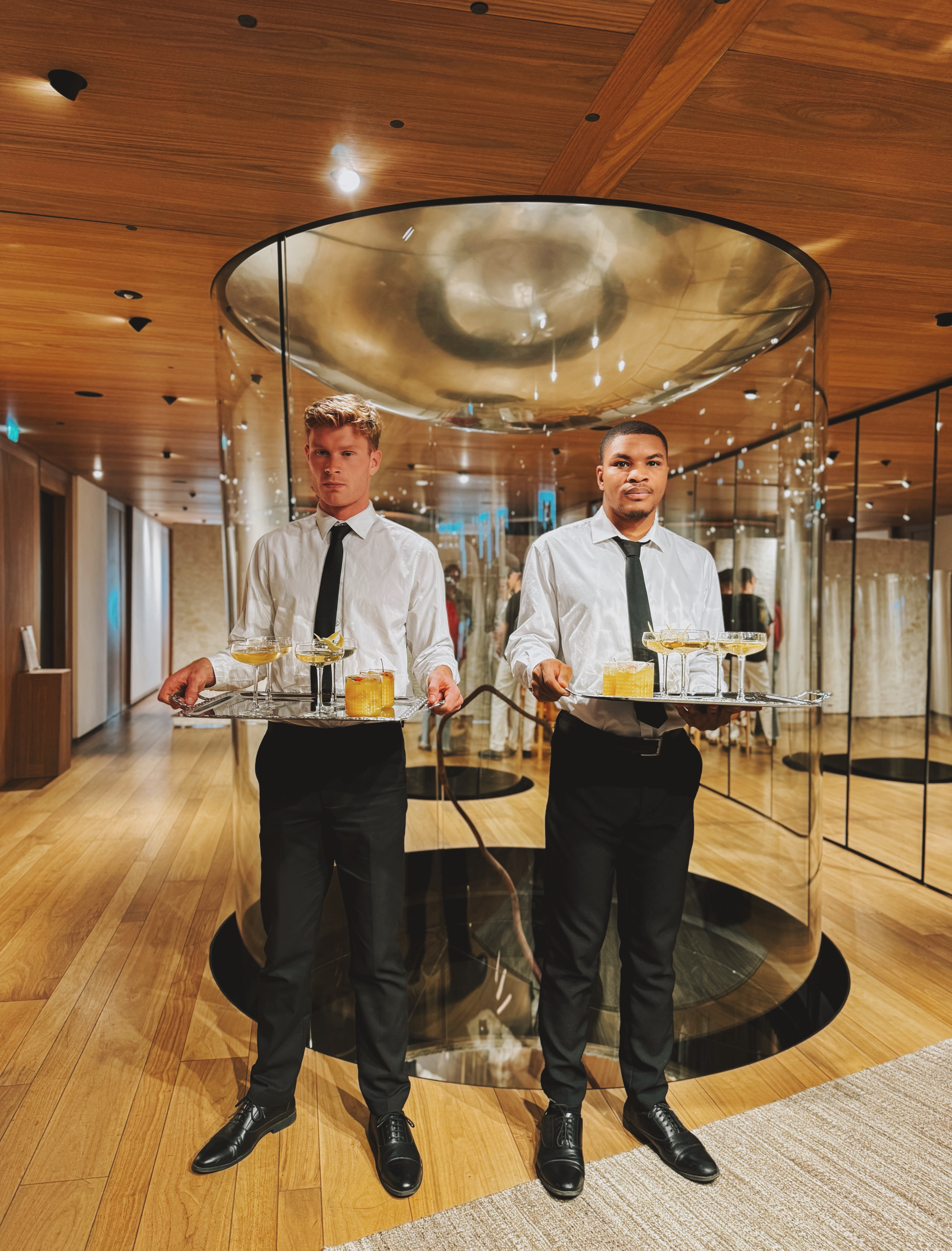 Two elegantly dressed servers stand in a modern venue with polished wood floors, showcasing artfully arranged cocktails in crystal glassware.