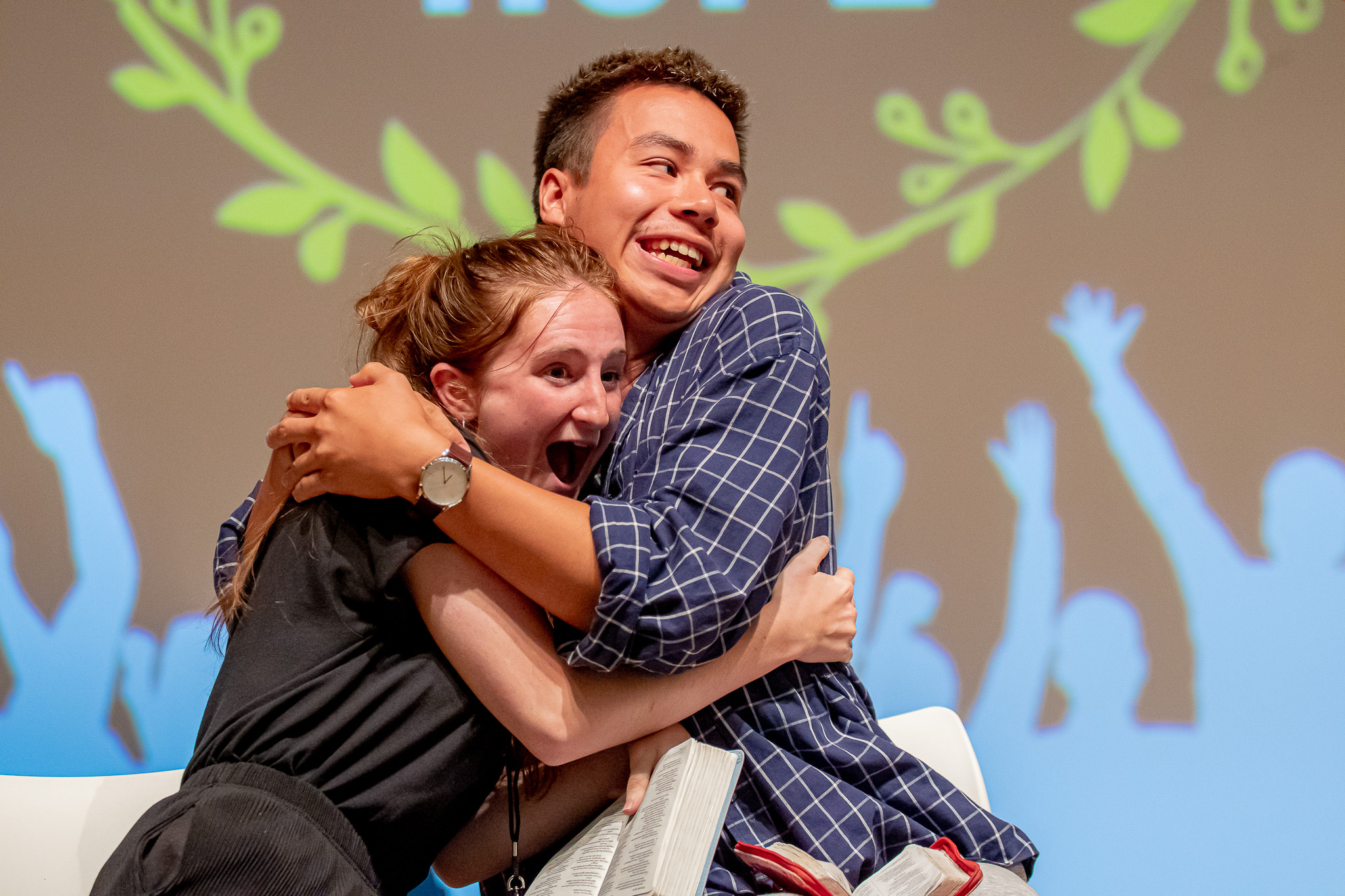 Two excited young people embrace on stage, celebrating joyfully, with an animated background featuring cheering silhouettes, conveying vibrant energy.