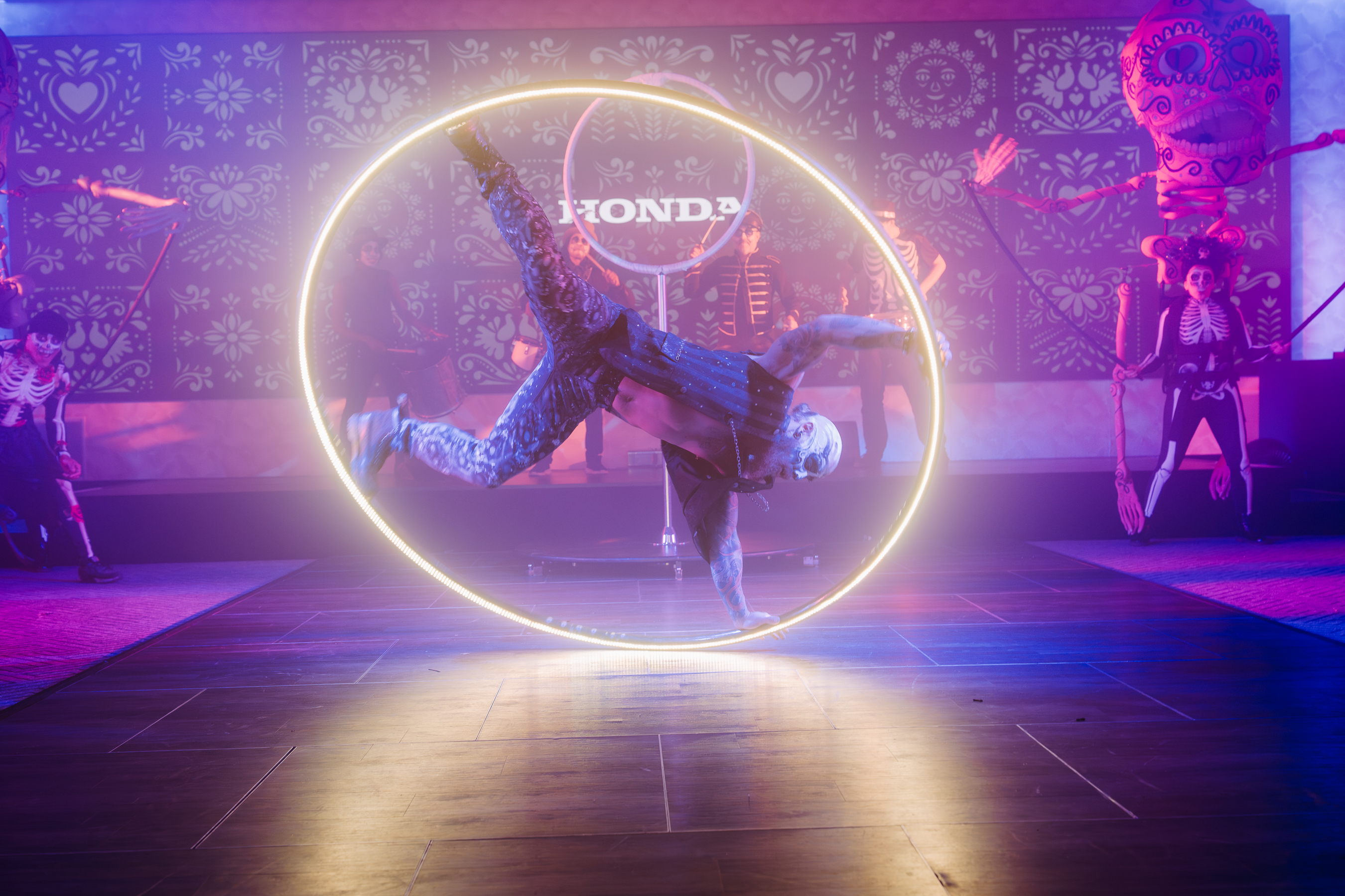 Neon-lit Honda event features a hoop acrobat center stage, with sugar-skull decor, purple haze, and polished floor.