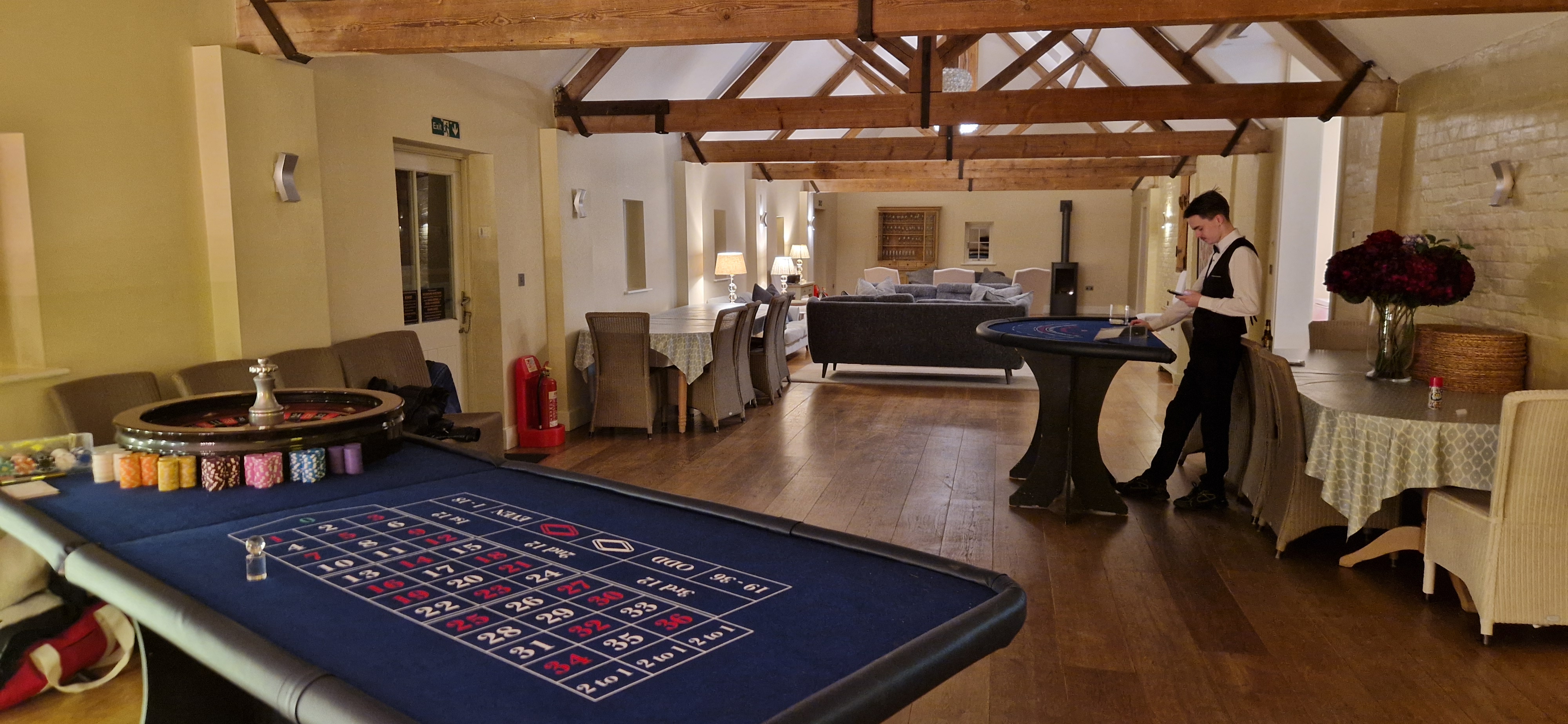 A cozy event space features dark wooden floors, elegant gray furniture, and two gaming tables under warm overhead lighting, creating a refined atmosphere.