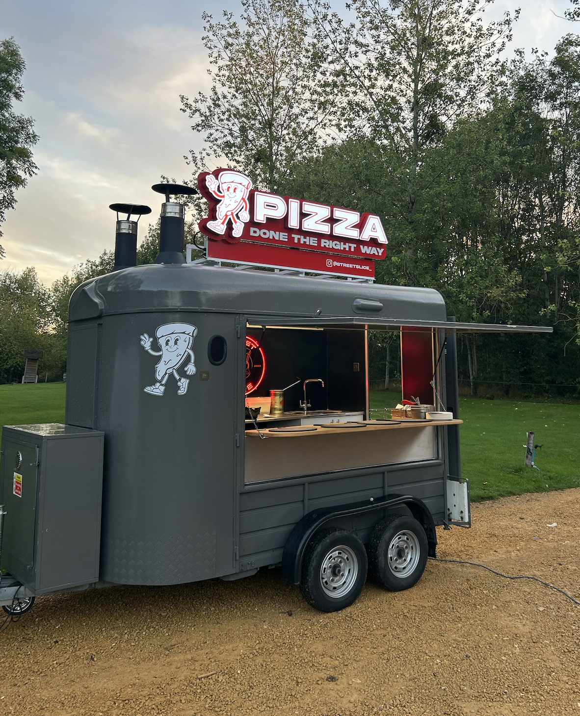 Gray pizza catering trailer with red neon sign, open service window, tidy wooden counter, and warm evening light on a grassy outdoor venue.