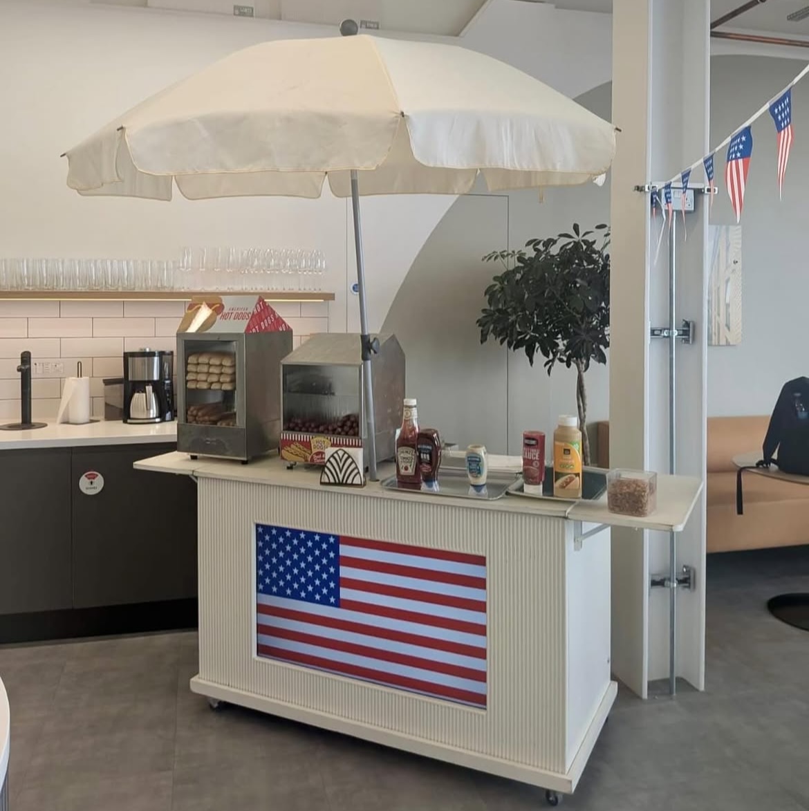 A charming hot dog cart with an American flag design, featuring neatly arranged condiments and buns, under a bright, cheerful umbrella.