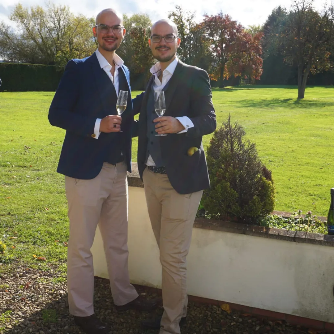 Two well-dressed men in tailored suits toast with champagne glasses against a lush green garden, radiating a cheerful, upscale atmosphere.