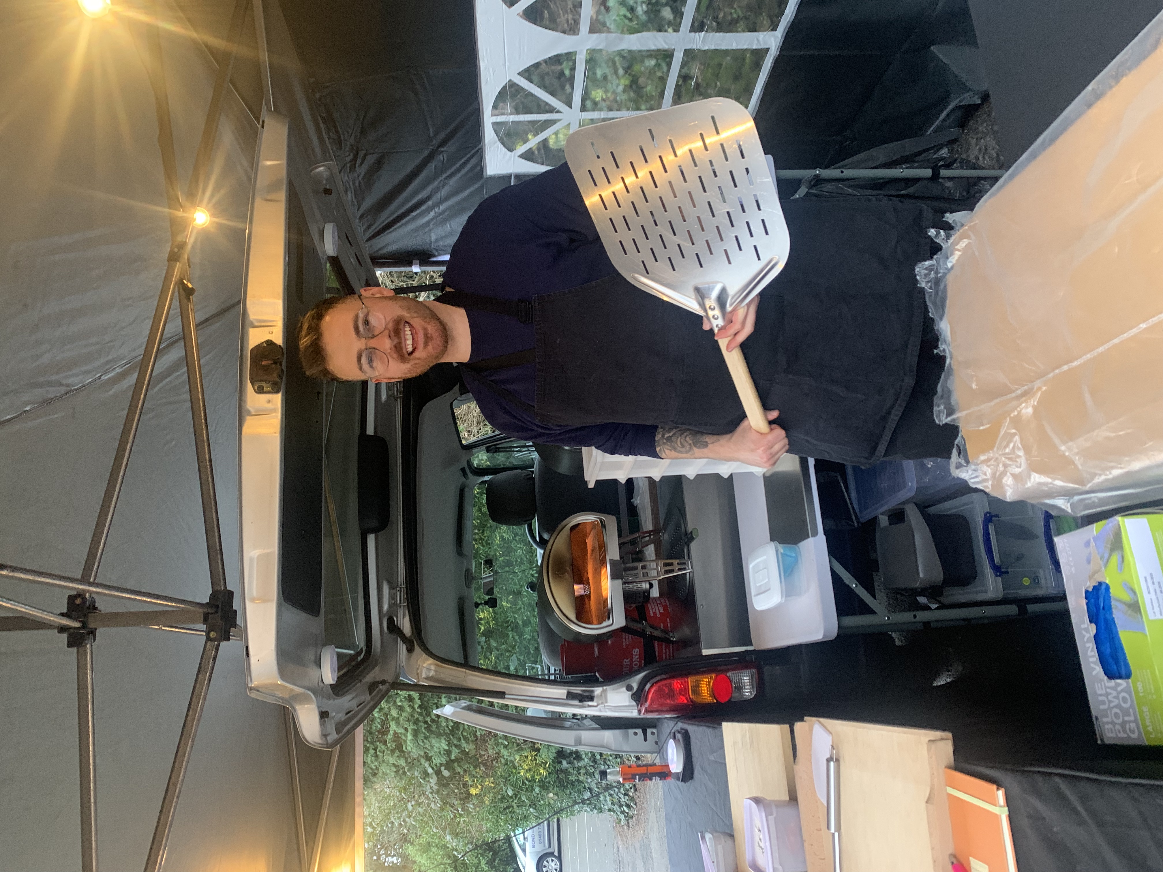 A cheerful chef stands under a tent, holding a metal pizza peel, surrounded by organized cooking equipment and plastic containers, evoking a welcoming catering vibe.