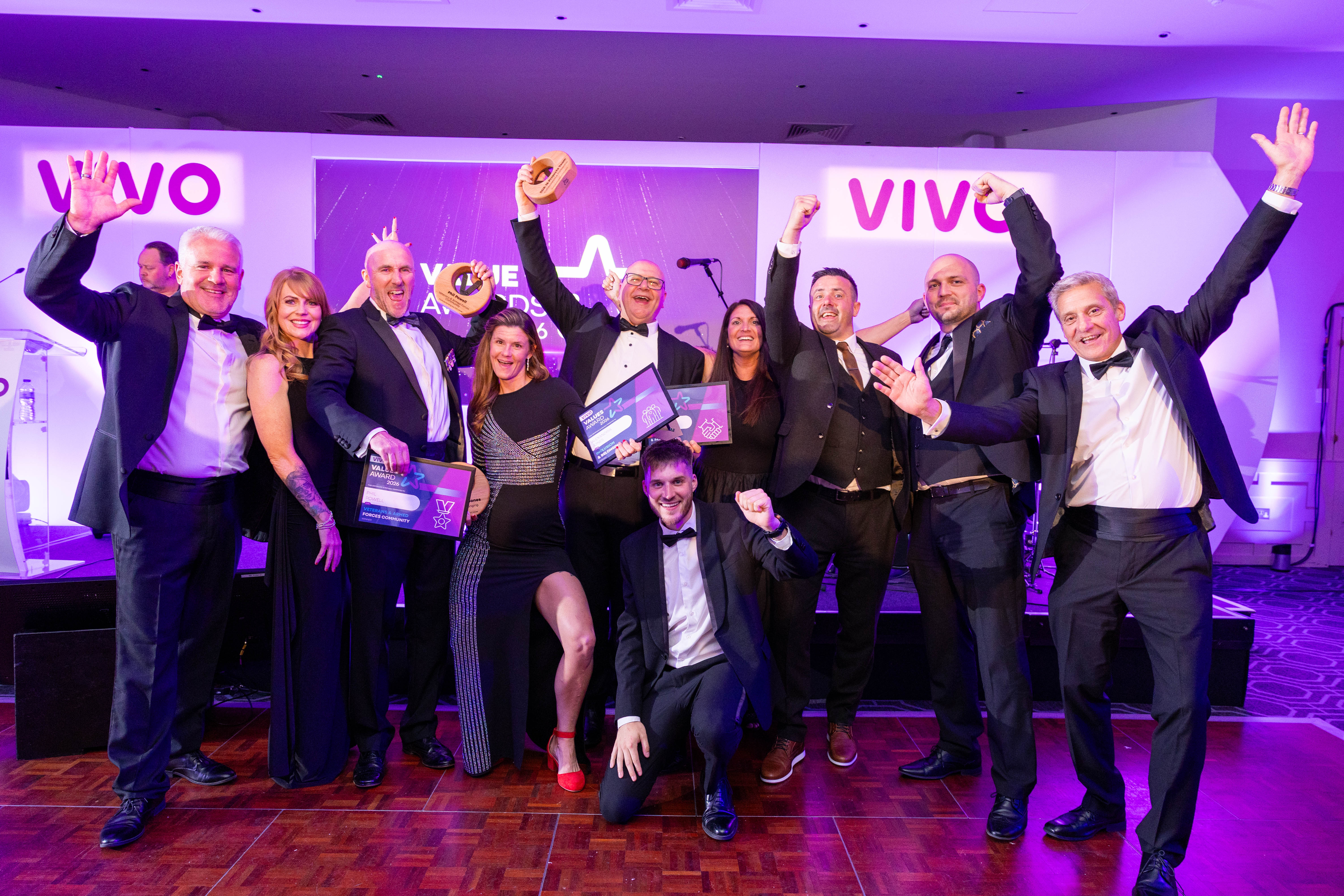 Cheering tuxedoed award winners pose on a purple-lit stage with VIVO branding, plaques, and trophies, capturing lively event catering energy.