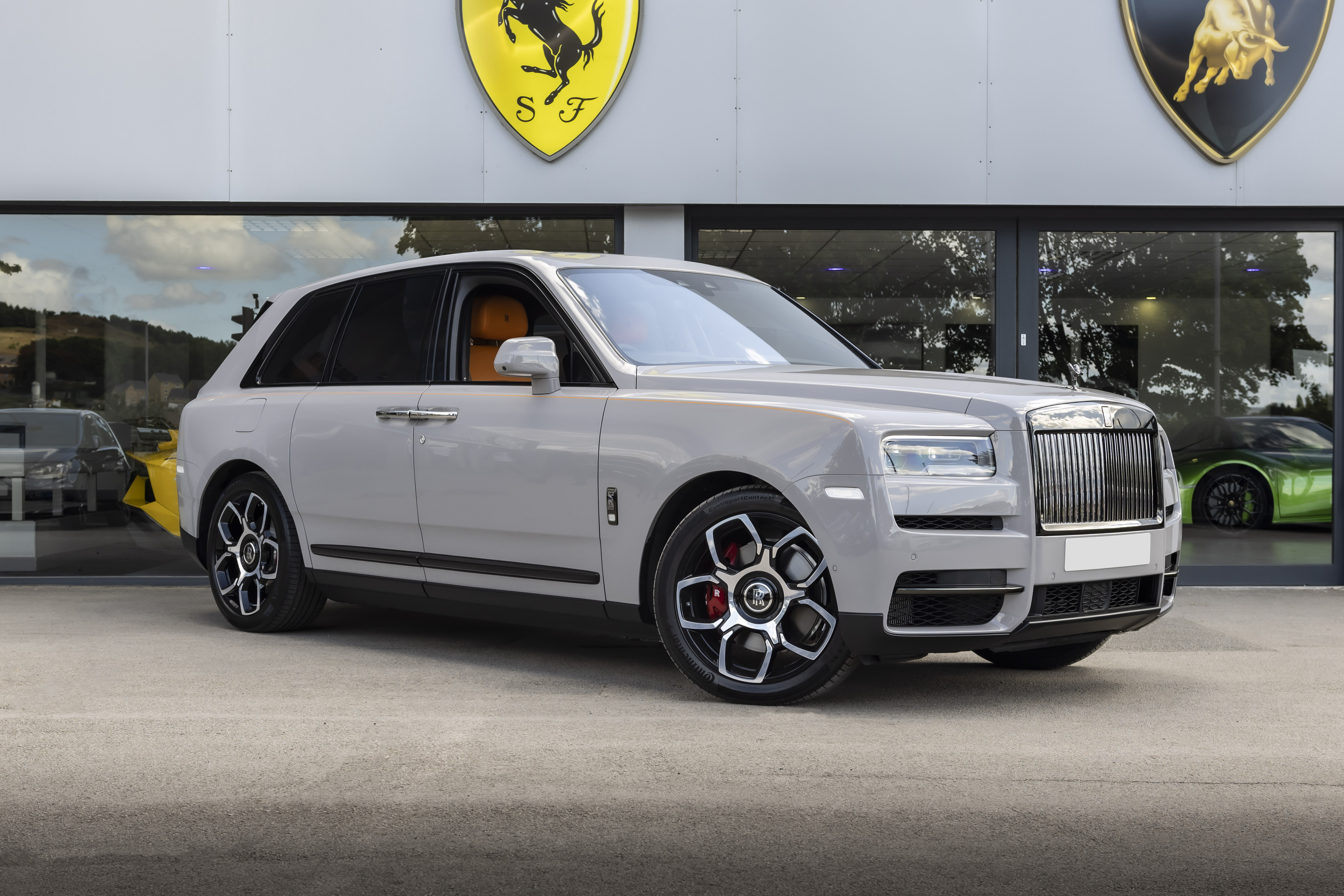 A sleek silver Rolls-Royce stands at a luxury dealership, with polished alloy wheels and a stunning interior reflecting vibrant orange accents.