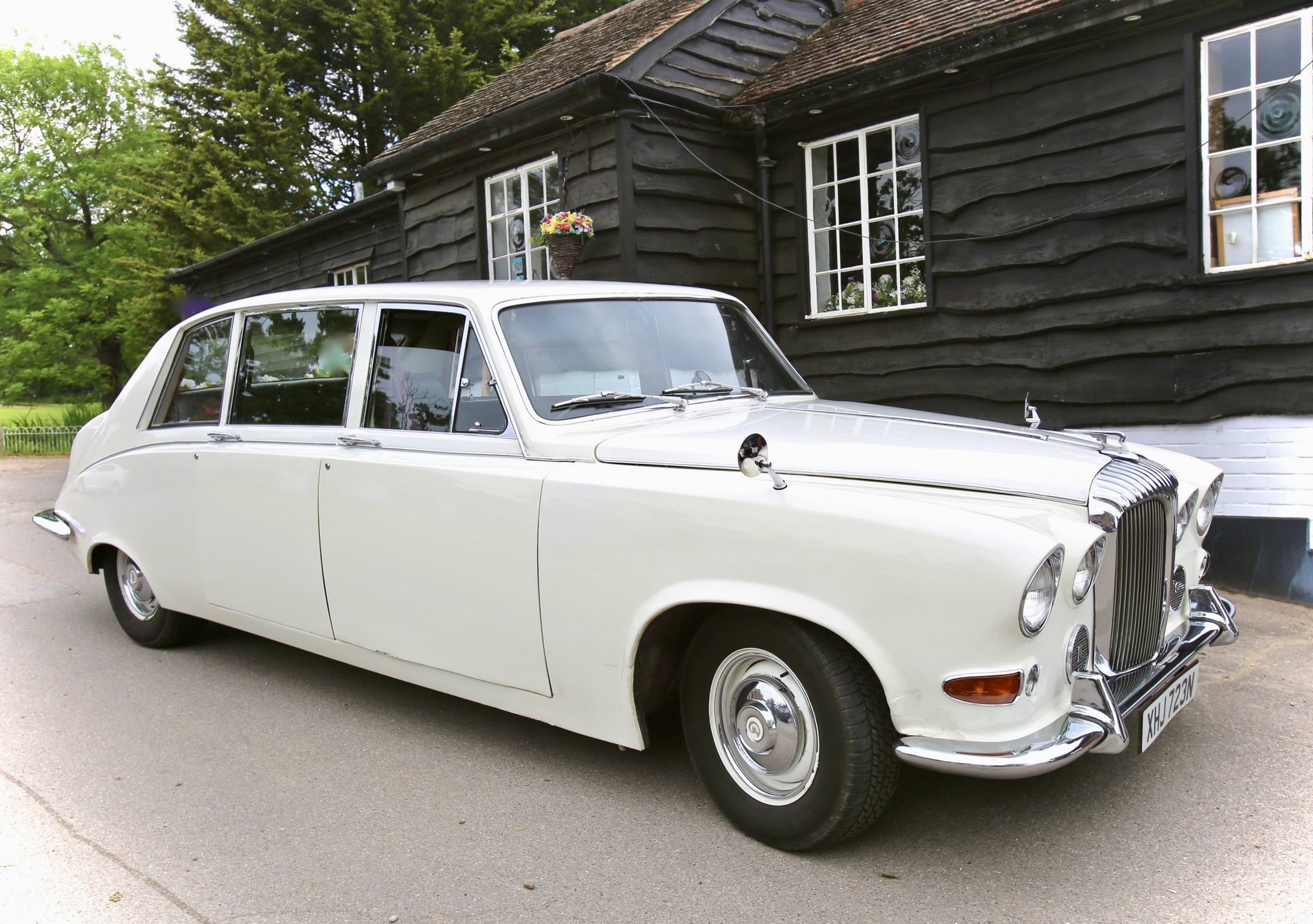 A classic white vintage car with chrome accents parked beside a rustic black wooden house, illuminated by soft daylight, exuding elegance.