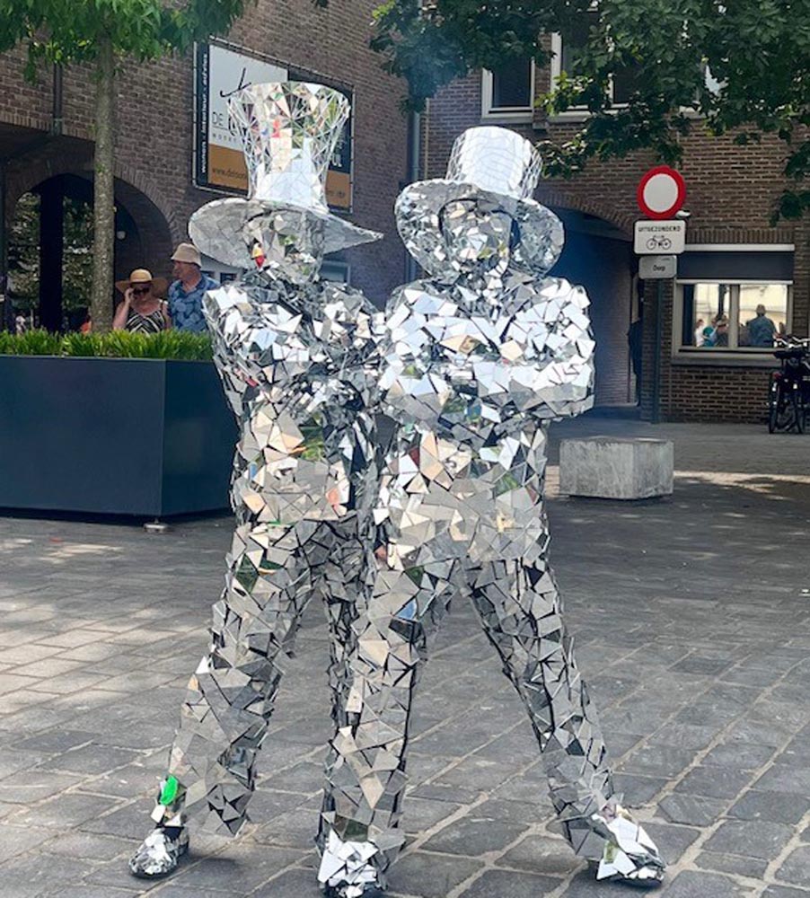 Two performers in dazzling mirrored costumes strike a pose in a sunlit square, surrounded by greenery and a rustic urban backdrop.