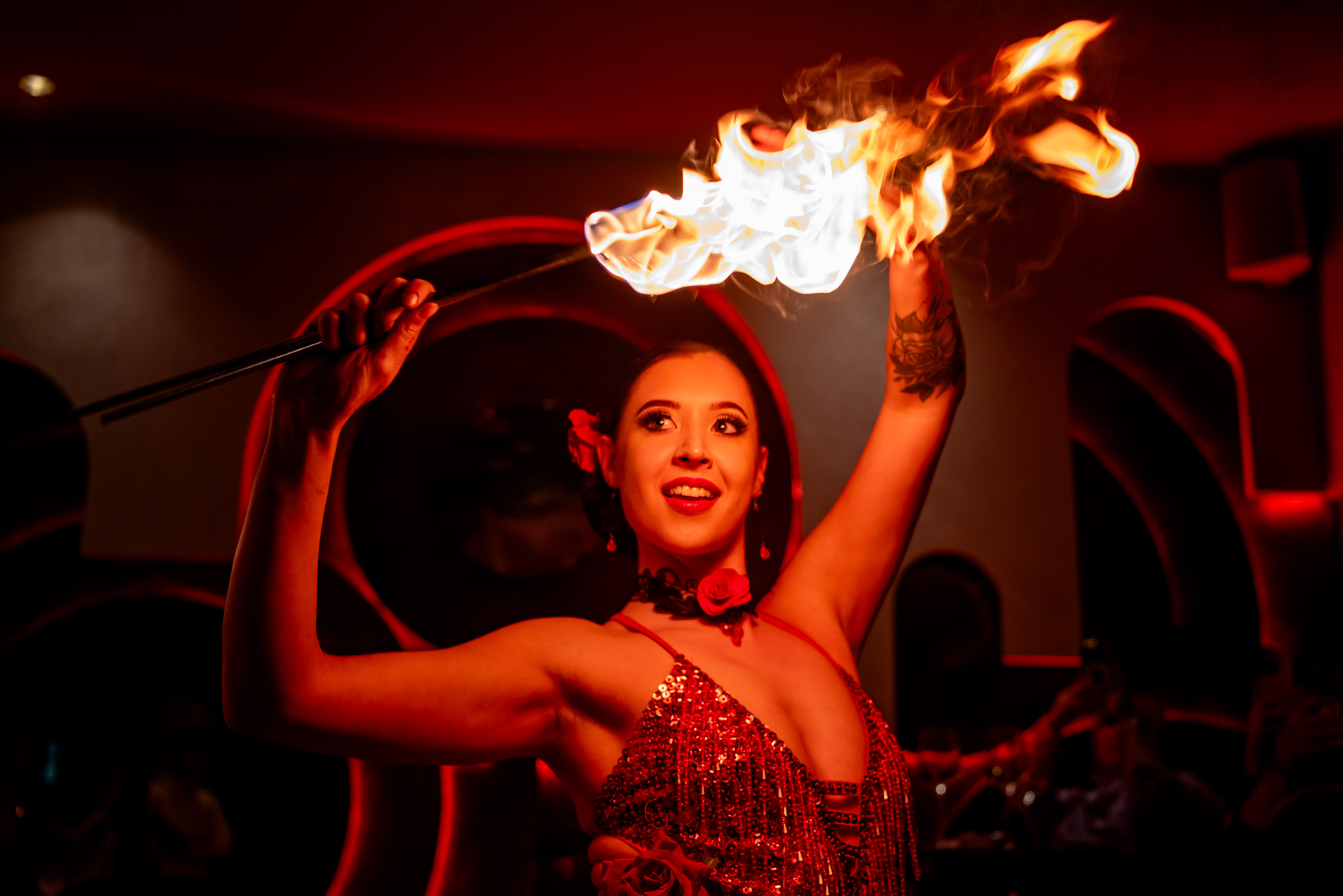 A performer in a shimmering red sequined dress emits flames, surrounded by warm red light. The intimate venue buzzes with energy, enhancing the captivating act.