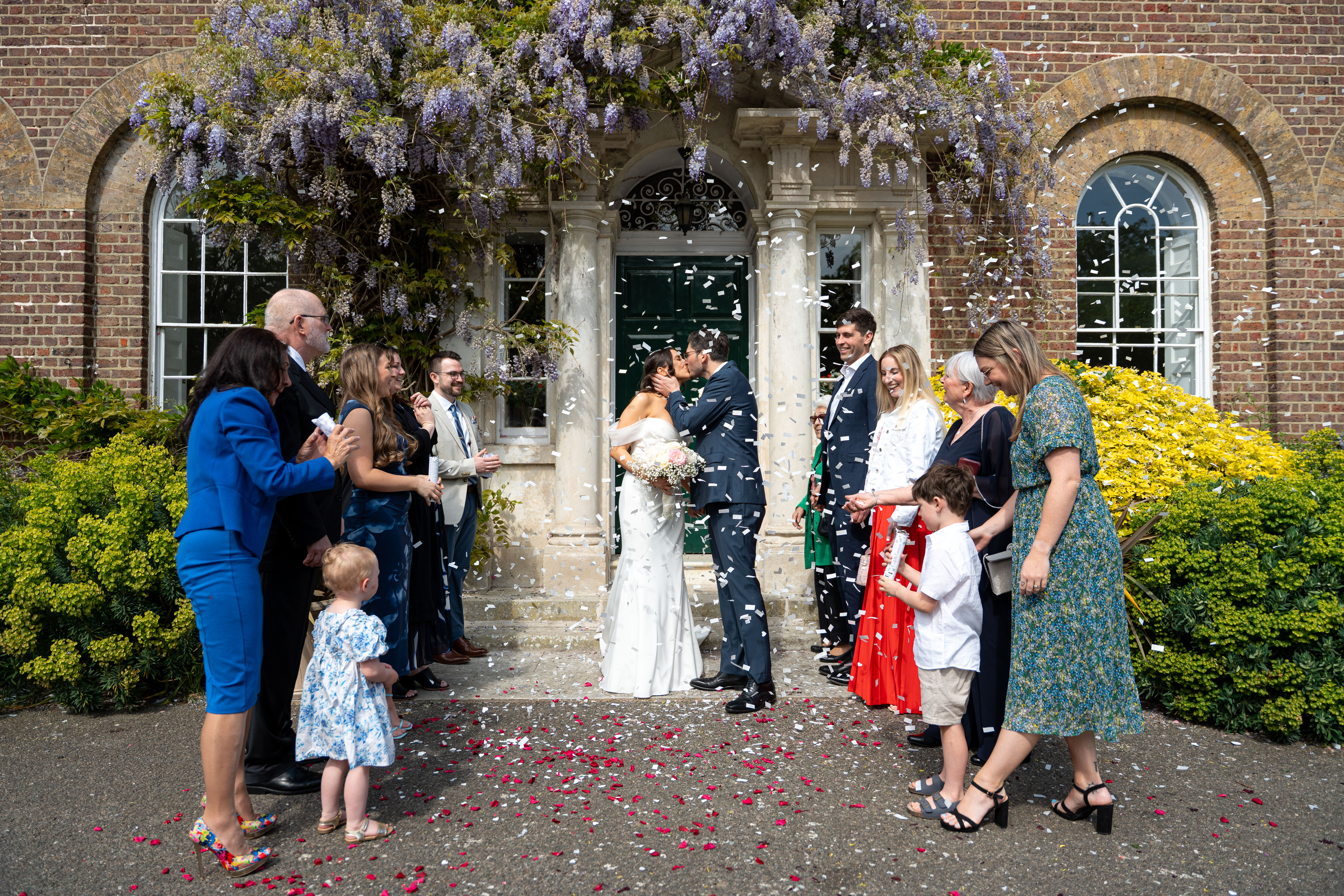 Bride and groom kiss beneath purple wisteria outside a brick manor, guests lining the aisle amid confetti, bright wedding-day atmosphere.