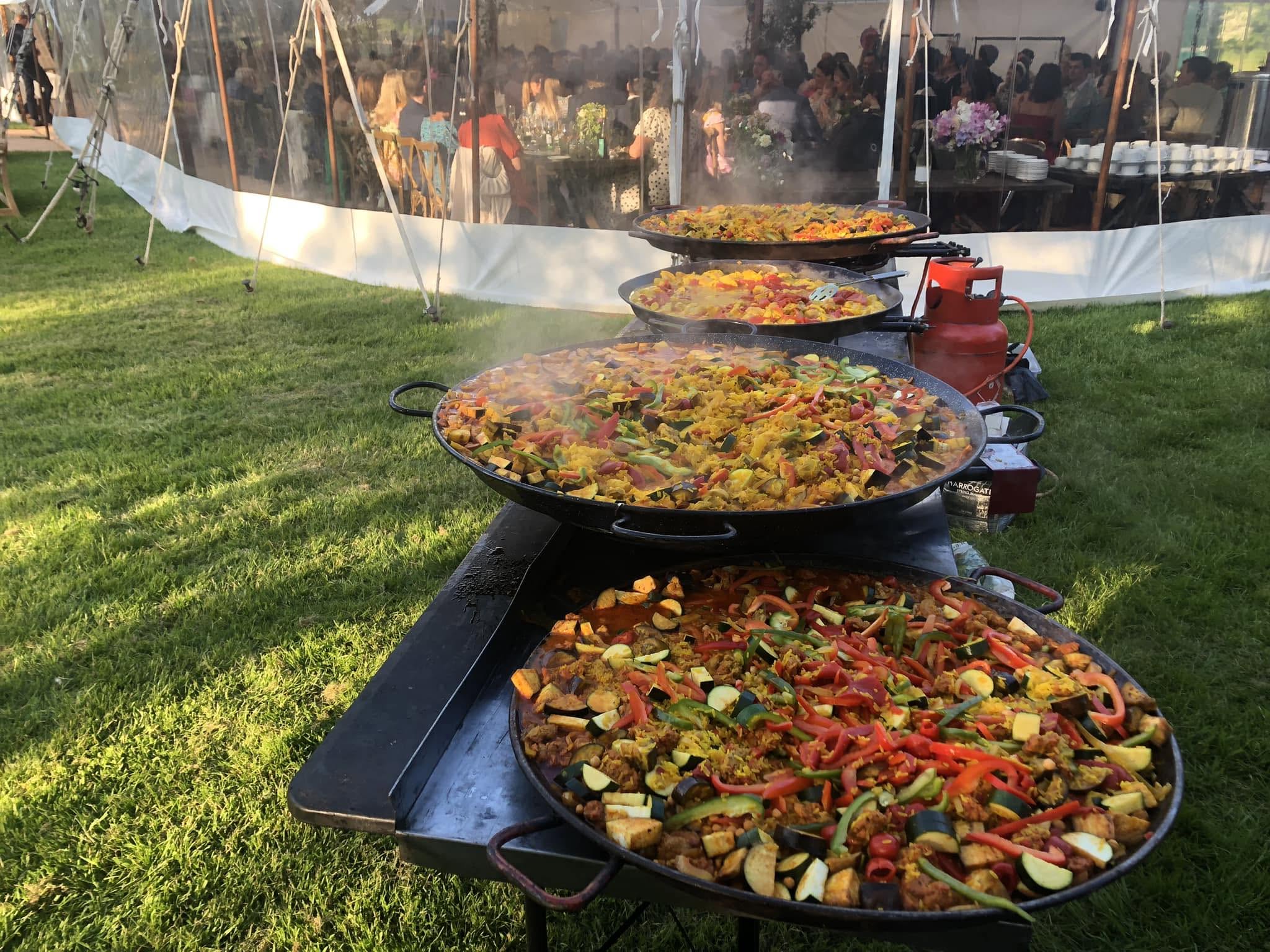 Outdoor catering buffet of steaming paella pans with colorful vegetables on black stands, beside a tented event in warm daylight.