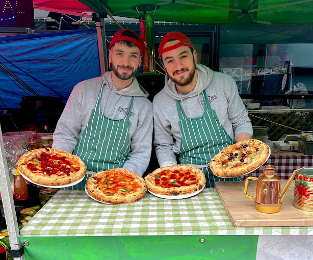 Two cheerful young men in striped aprons proudly display four artisanal pizzas on a green-checkered table, complemented by a vintage copper pot.