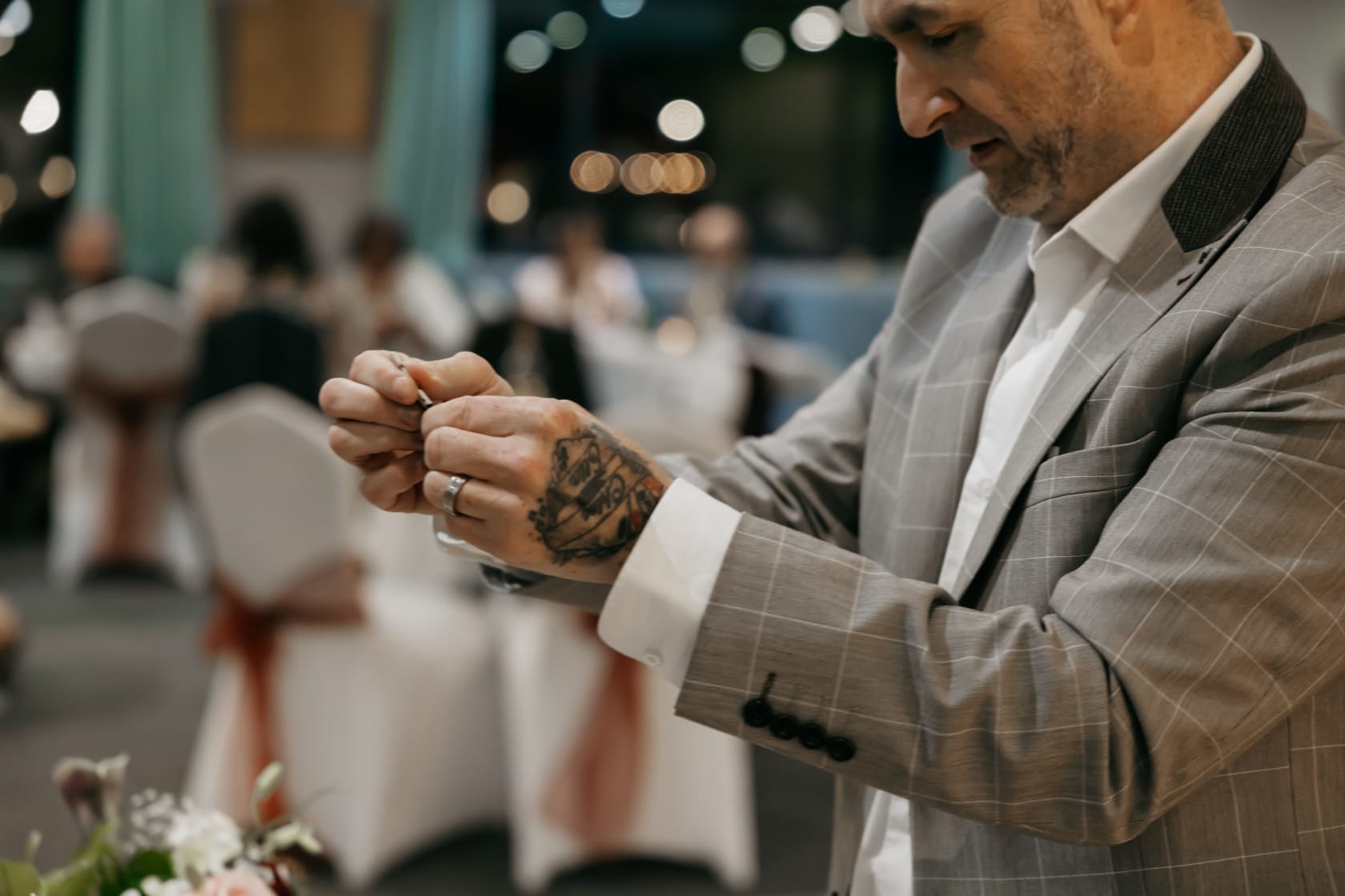 Tattooed man in a gray plaid suit adjusts a detail at a wedding reception, with blurred white chair covers, soft bokeh, and warm ambient catering lighting.