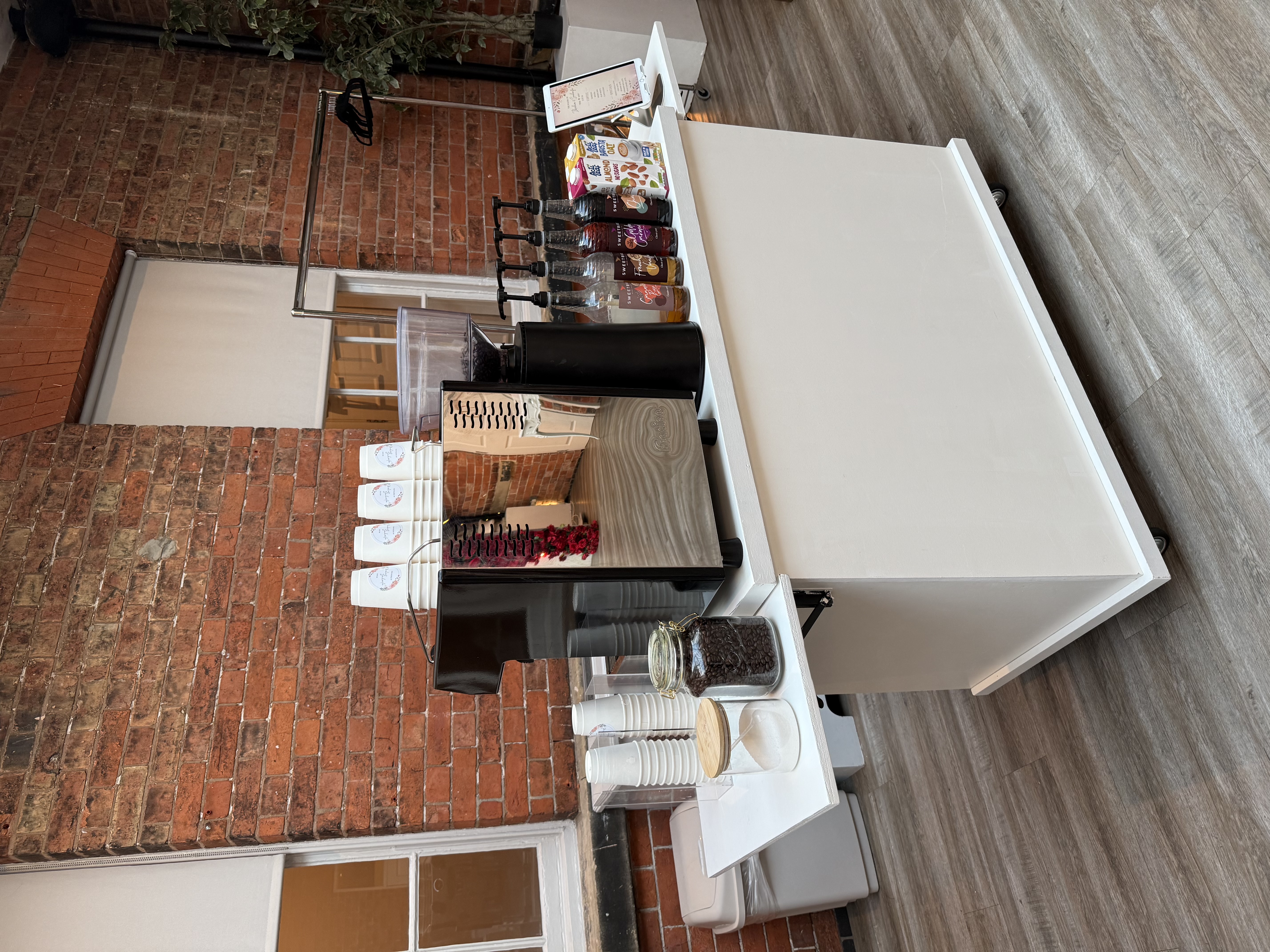 Elegant coffee catering cart with espresso machine, syrup bottles, oat milk, stacked cups, and polished glass jars in a bright brick venue.