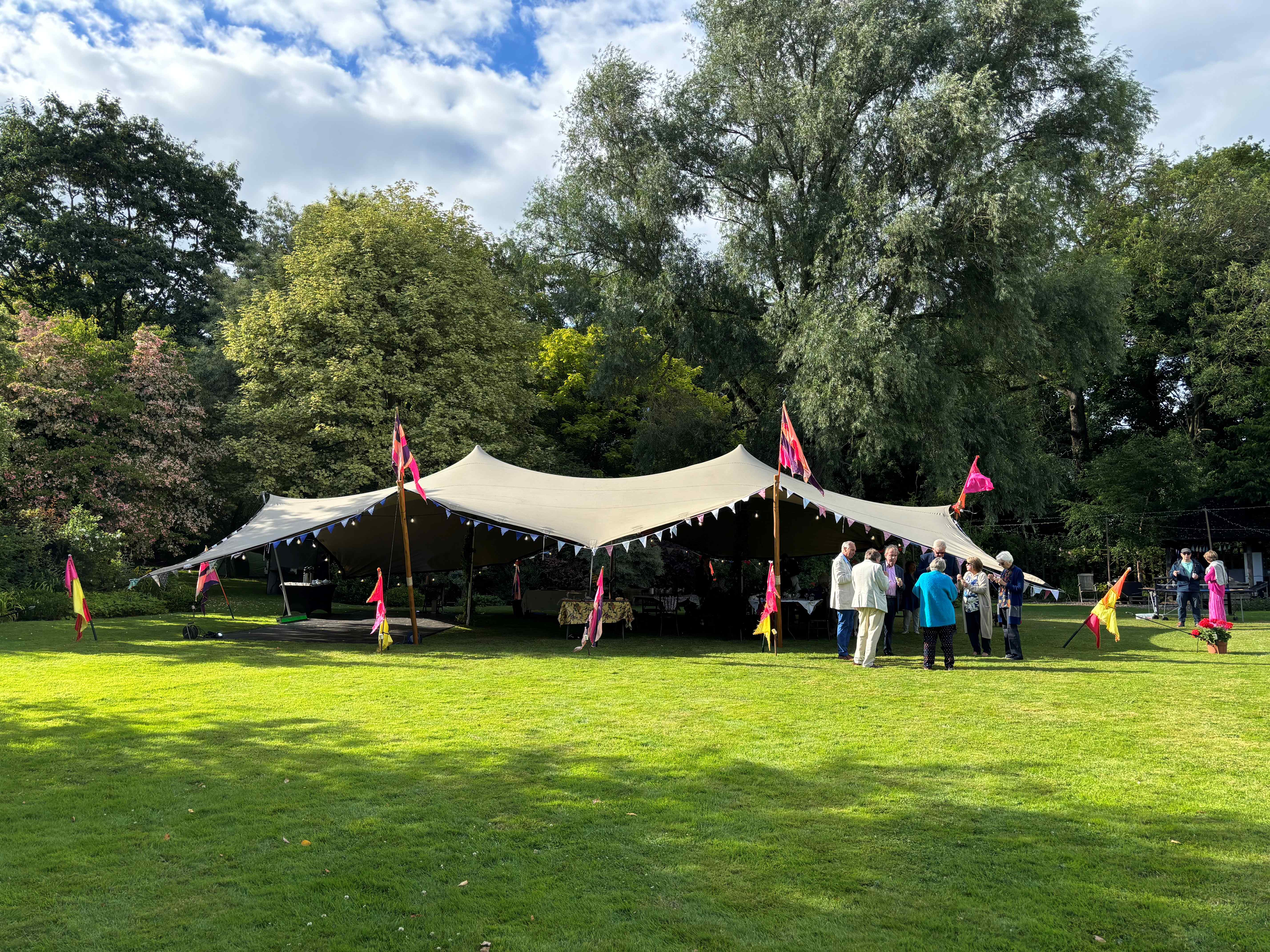 A vibrant marquee tent adorned with colorful flags hosts a cheerful gathering on bright green grass, surrounded by lush trees and joyous conversations.