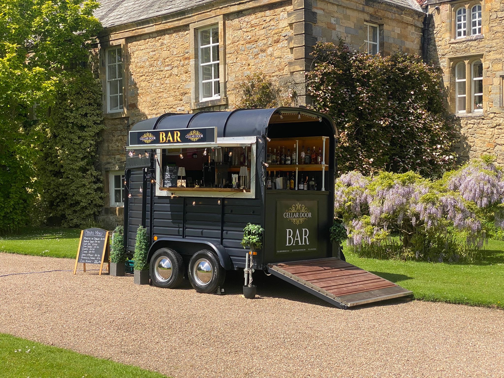A vintage black mobile bar, "Prosecco Palace," features polished glassware and potted greenery, exuding a charming, inviting atmosphere in daylight.