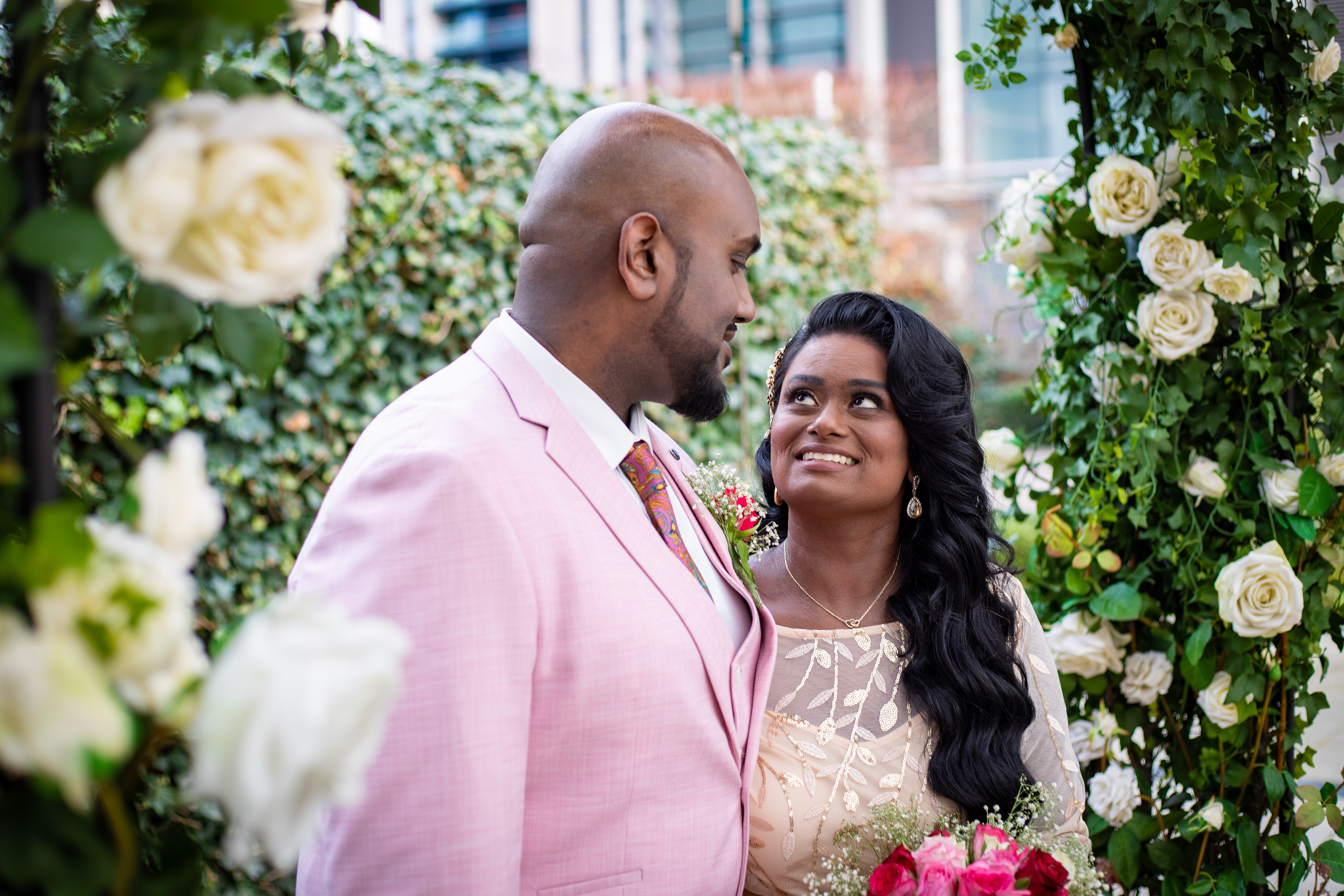 A stylish couple sits at a floral-adorned table, surrounded by elegantly dressed guests, in bright, inviting lighting, exuding joyful celebration.