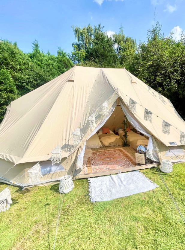 A cozy bell tent with soft cushions and a vibrant patterned rug invites relaxation under bright sunlight, enhancing a glamping experience.