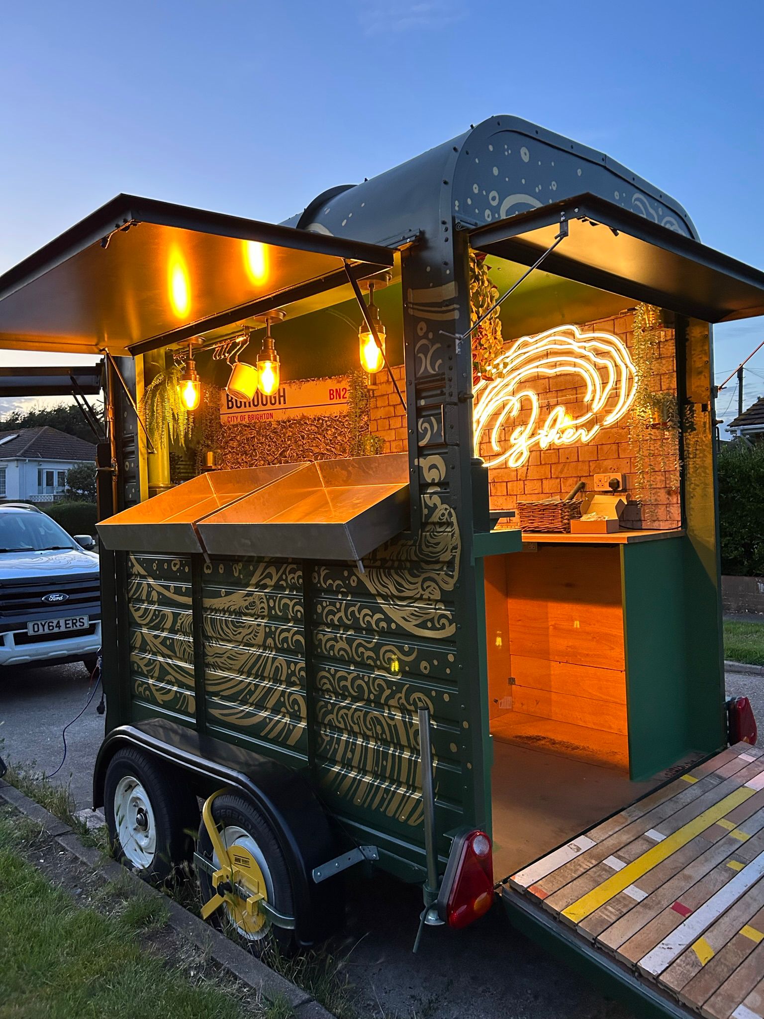 A vintage catering van features a charming wooden deck, warm pendant lighting, and vibrant green walls, creating a cozy atmosphere.