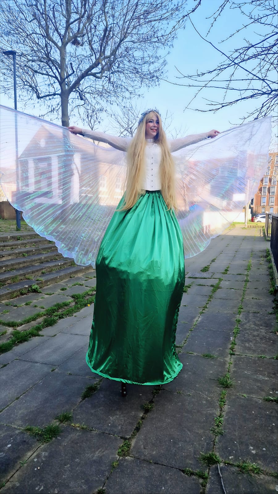 A performer in a flowing green satin skirt and white top, with translucent wings, stands on stone steps under a clear sky, exuding whimsy.