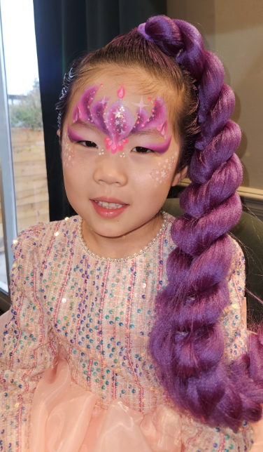 Young girl in a sparkly pastel dress with purple braided wig and glitter face paint, lit by soft indoor daylight.