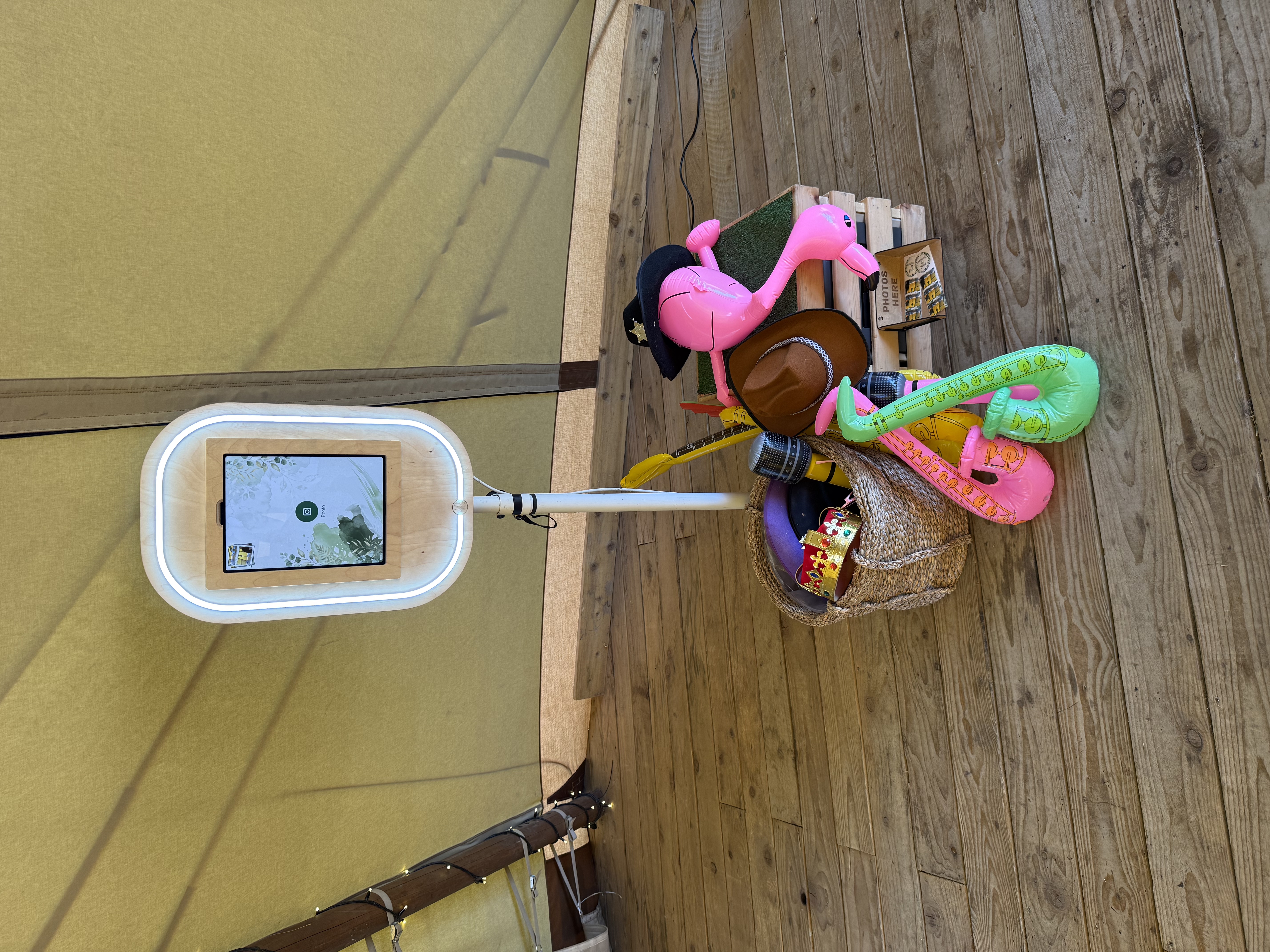 Photo booth kiosk with glowing white ring light on a rustic wood floor, flanked by pink flamingos, hats, props, and bright daylight.