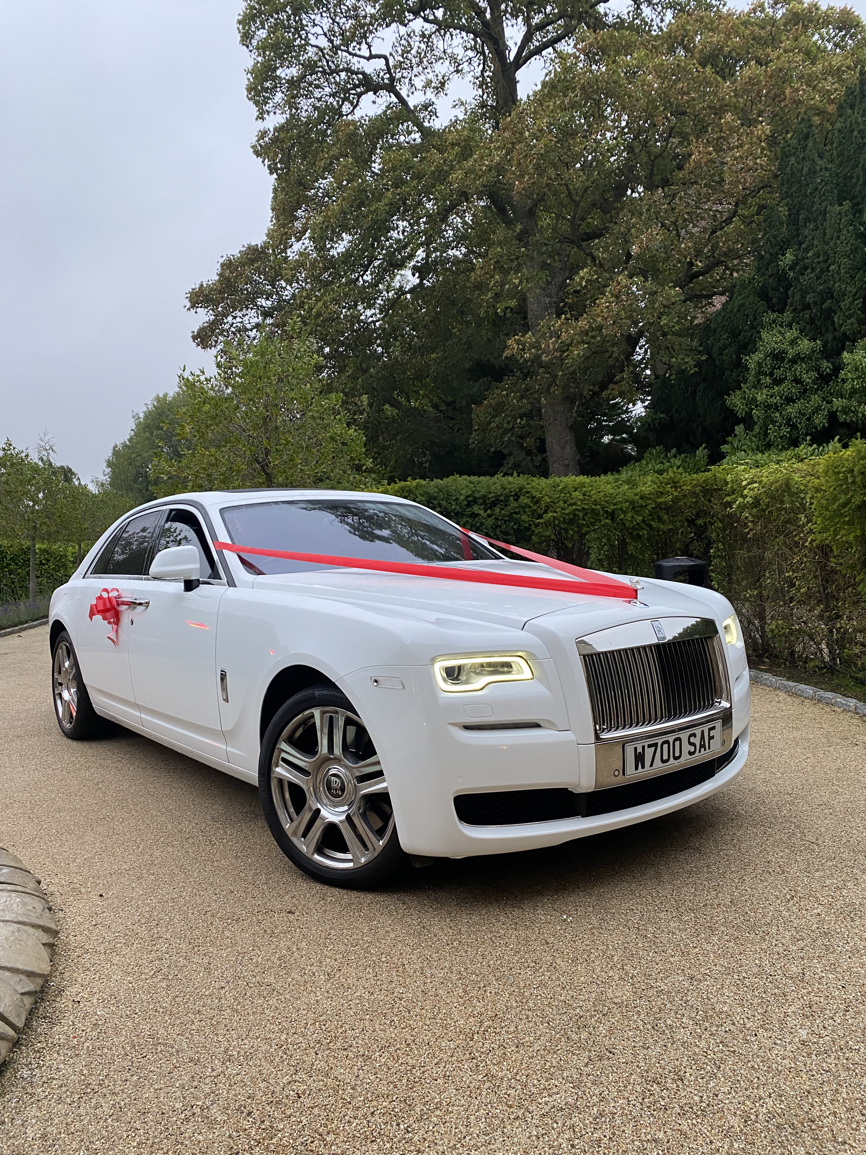 A pristine white Rolls-Royce adorned with a vibrant red ribbon is parked on a textured gravel driveway, surrounded by lush greenery.