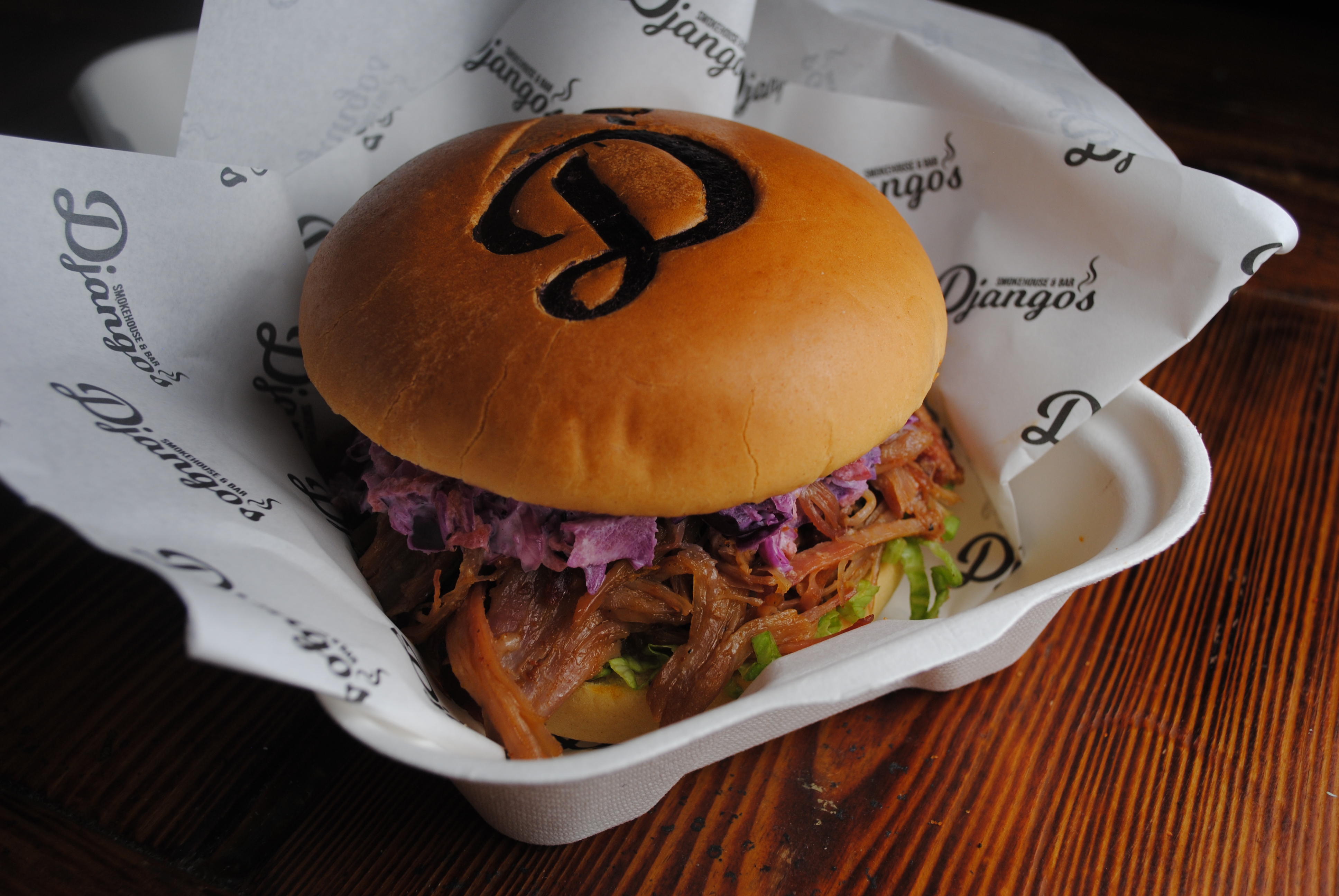 Gourmet pulled-pork slider with purple slaw in branded paper tray on a dark wood table, softly lit for polished catering presentation.