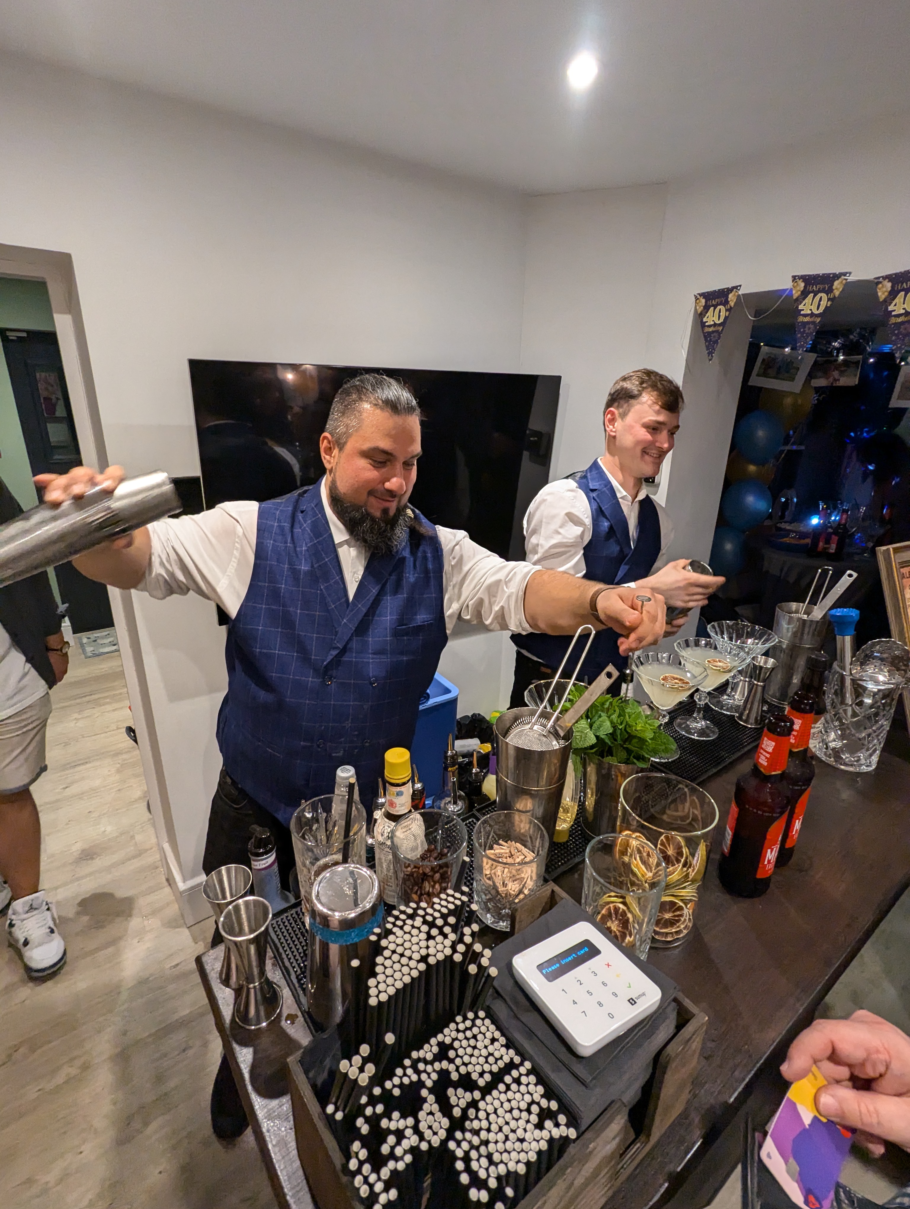 Two skilled bartenders in crisp blue vests craft cocktails amid polished glassware, fresh mint, and stylish utensils, enhancing the festive vibe.