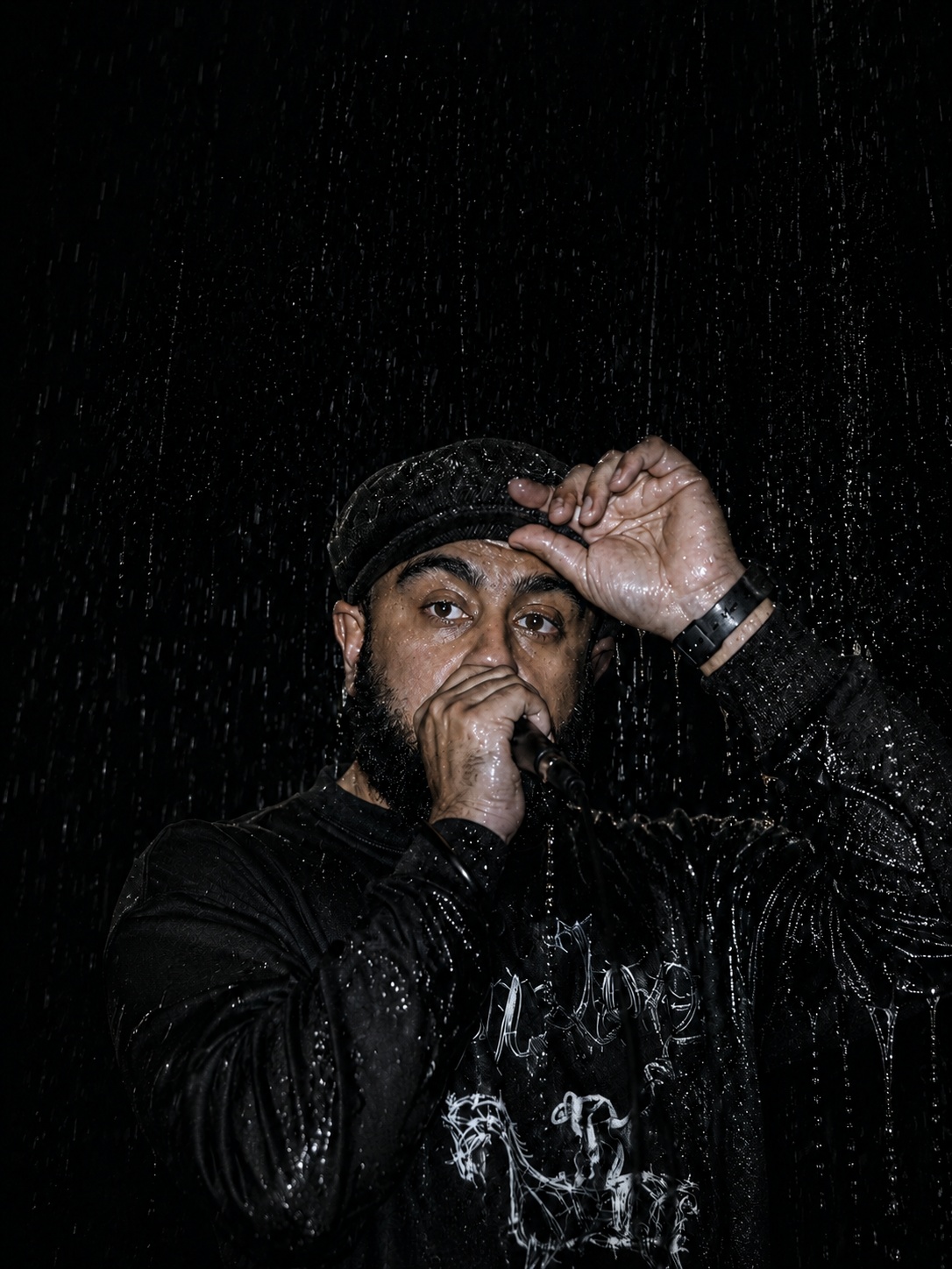Rain-soaked live performer in black cap and graphic shirt sings into a mic, spotlighted against a dark, moody concert stage.