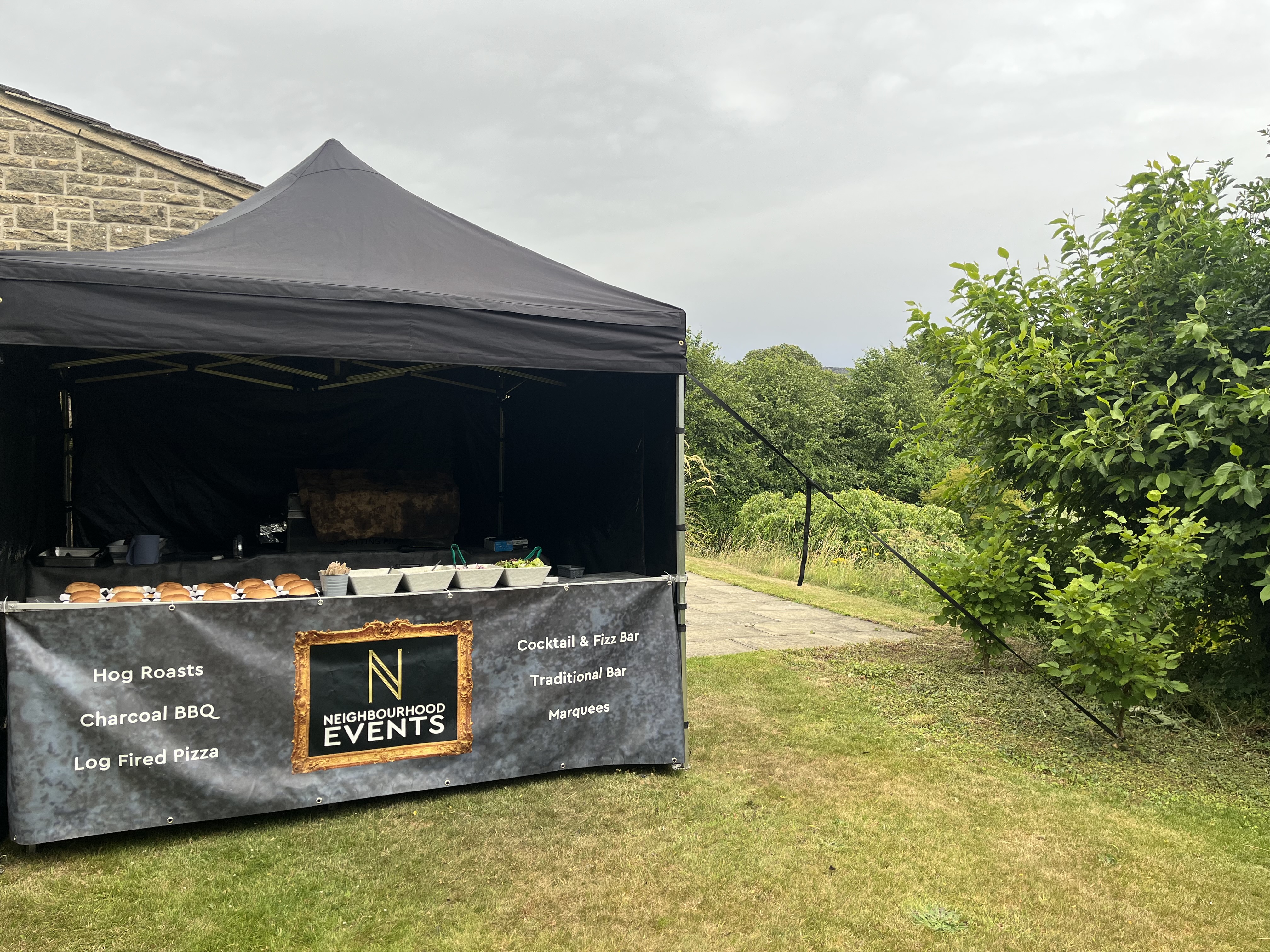 A sleek black tent showcases a buffet with neatly arranged hog roast buns and stainless trays, set against a lush green backdrop.