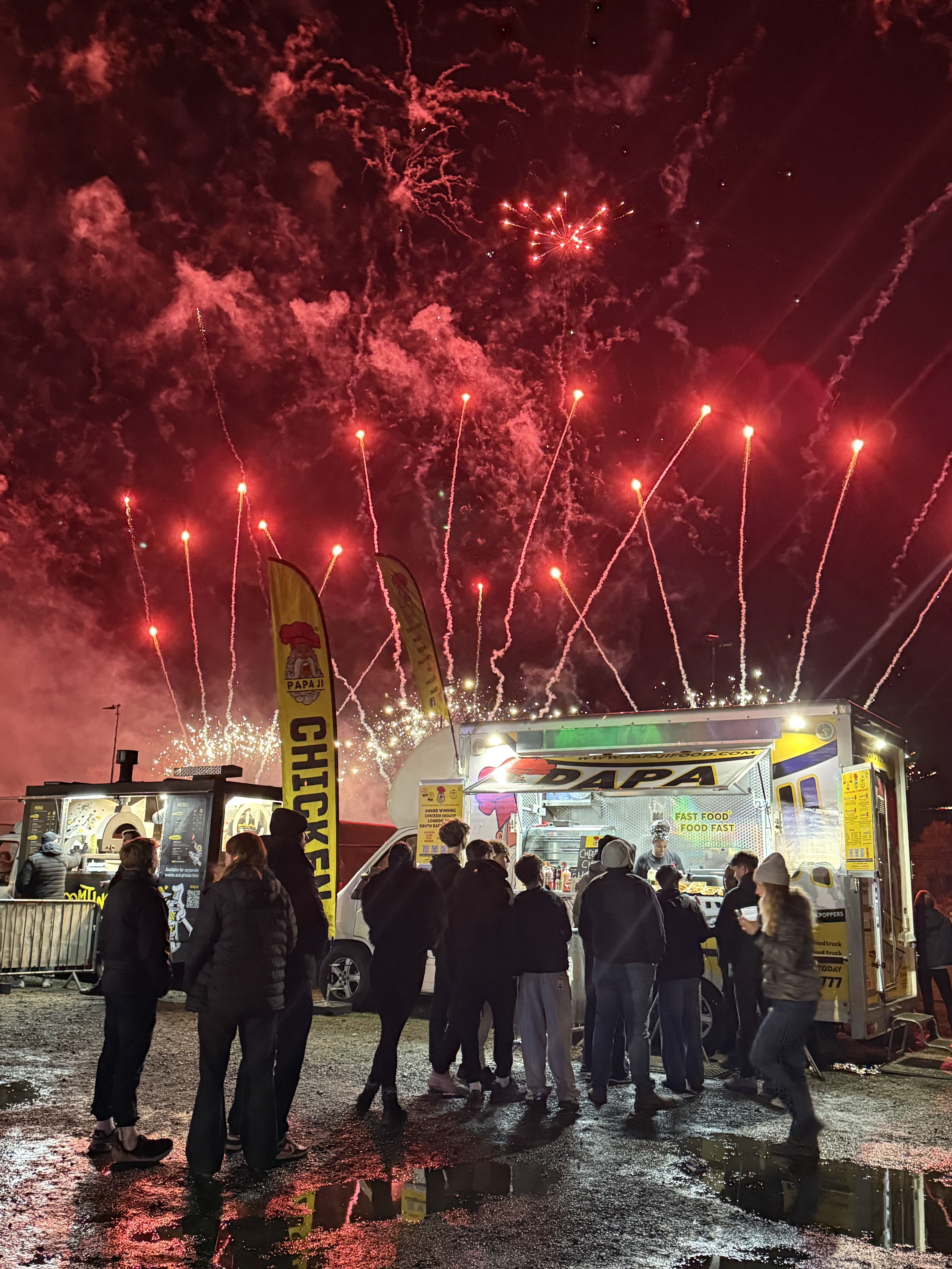 A bustling food truck serves late-night snacks under vibrant red fireworks, with a crowd in cozy jackets, reflecting excitement and warmth.