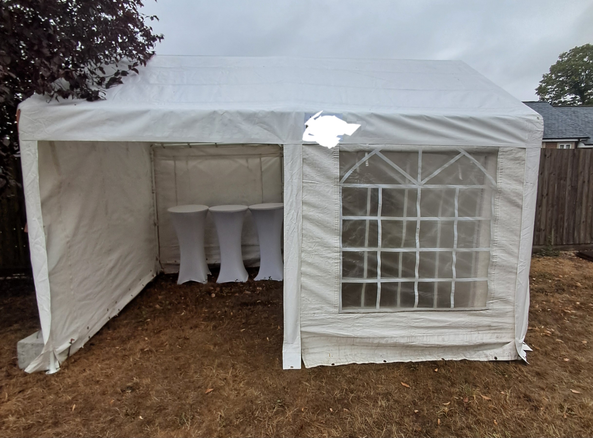 White marquee tent with clear window panel and three cocktail tables on dry grass, clean catering setup under gray daylight.