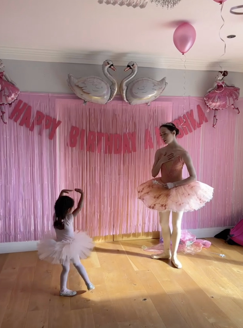 A whimsical birthday scene features two girls in ballet attire, surrounded by pink fringing and ballet-themed decorations, celebrating joyfully.