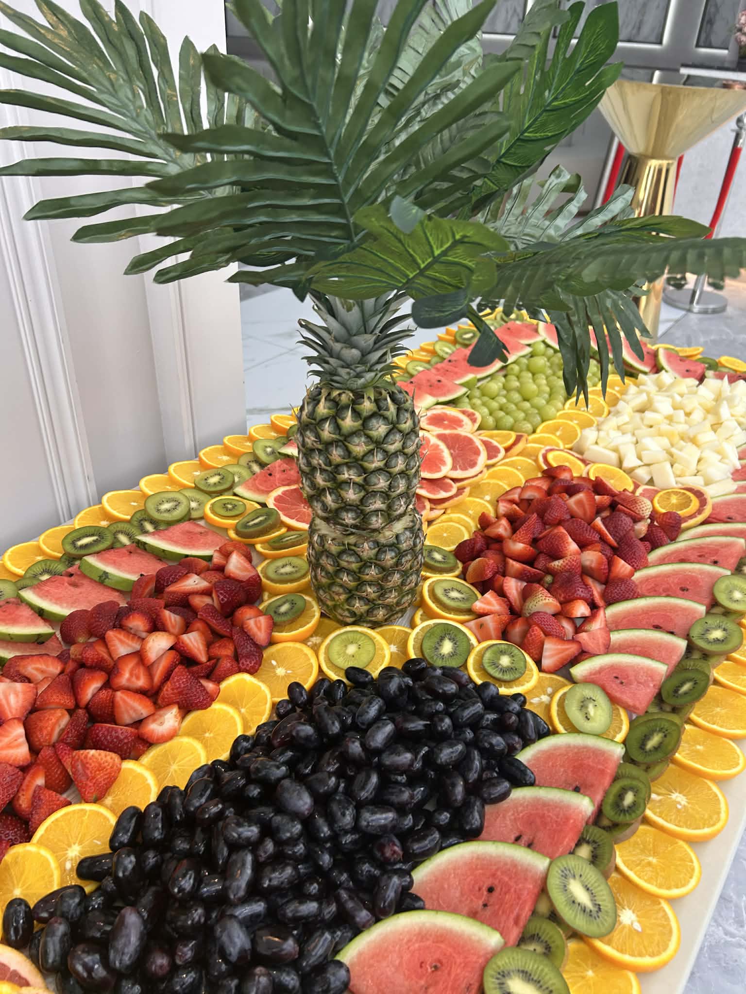 A vibrant fruit display features watermelon, strawberries, oranges, and grapes arranged artistically around a central pineapple, creating a lively buffet atmosphere perfect for catering events.