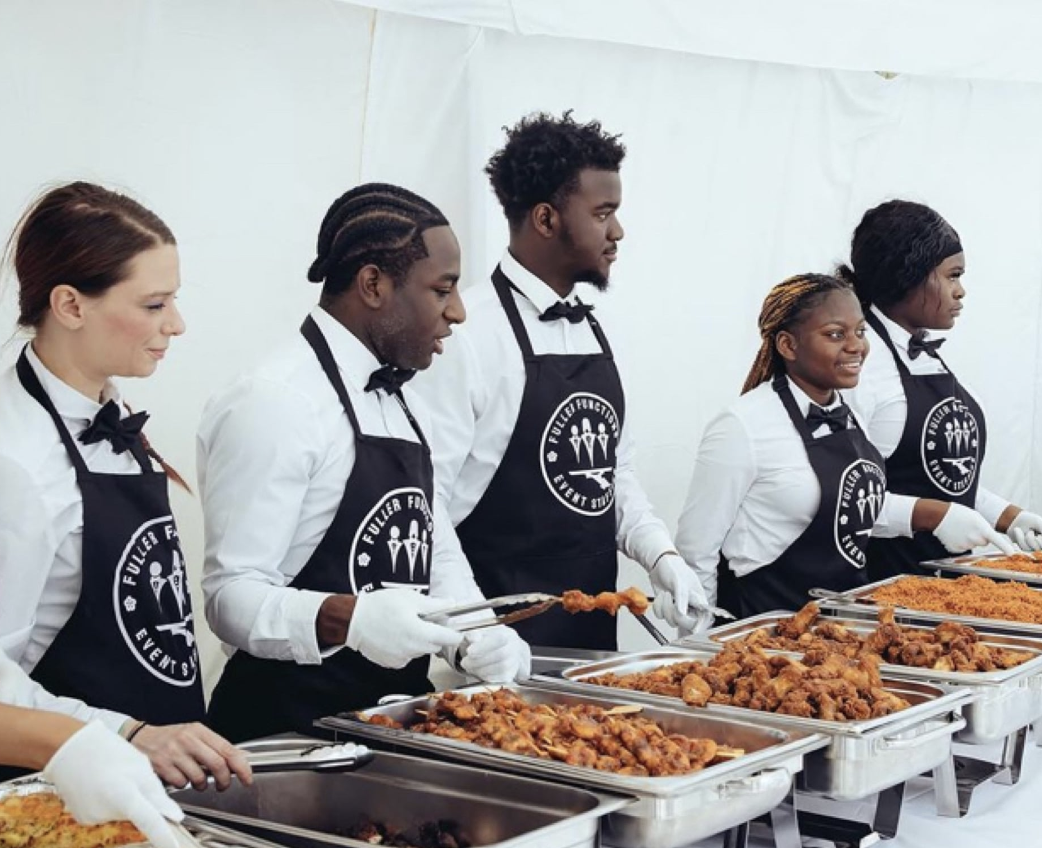 Attentive staff in black aprons serve a vibrant buffet of savory dishes, amid bright, inviting tent lighting for a polished catering experience.