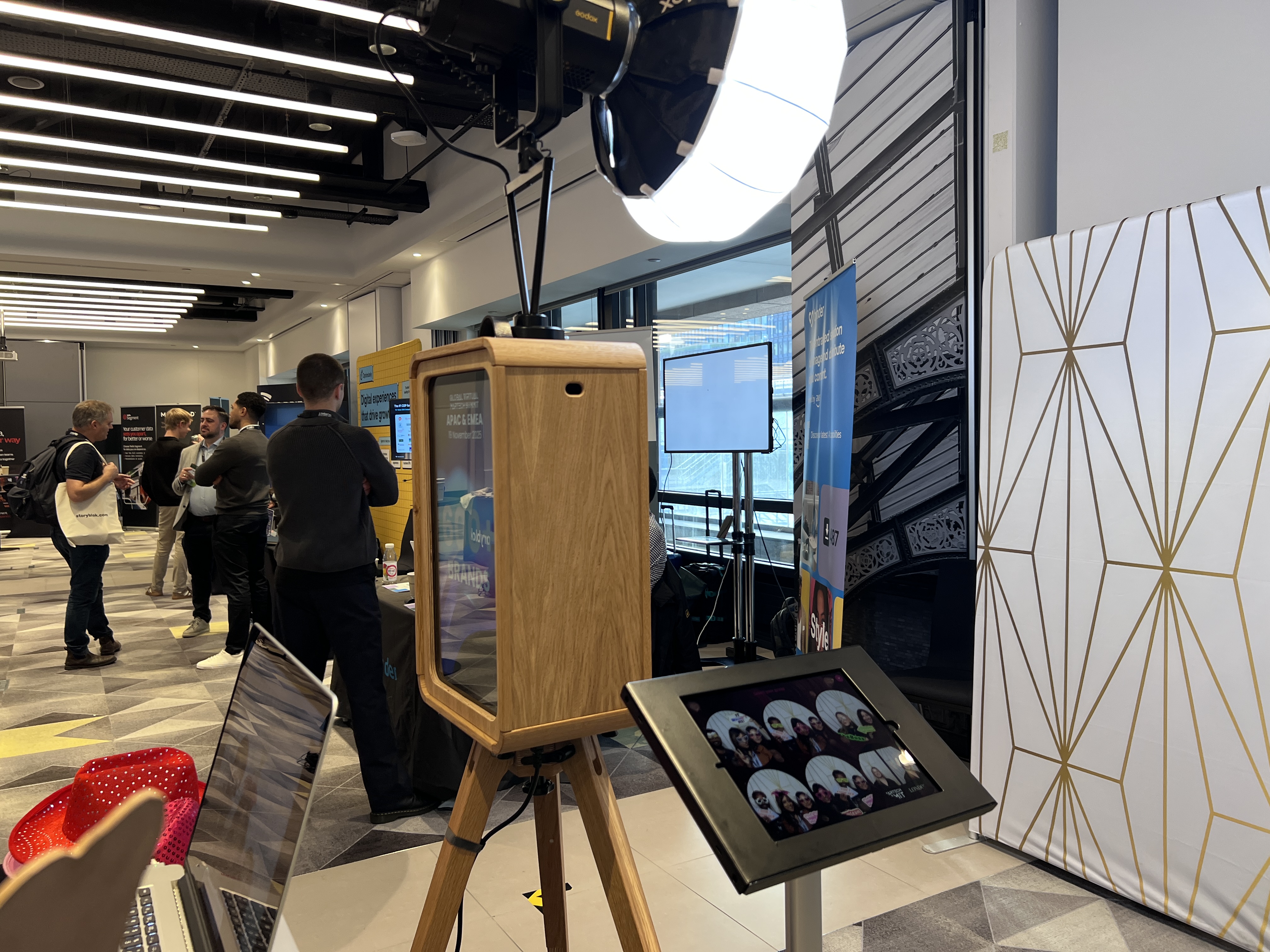 A modern event space showcases a sleek wooden kiosk, attendees engage in conversations, illuminated by bright overhead lights, creating a lively atmosphere.