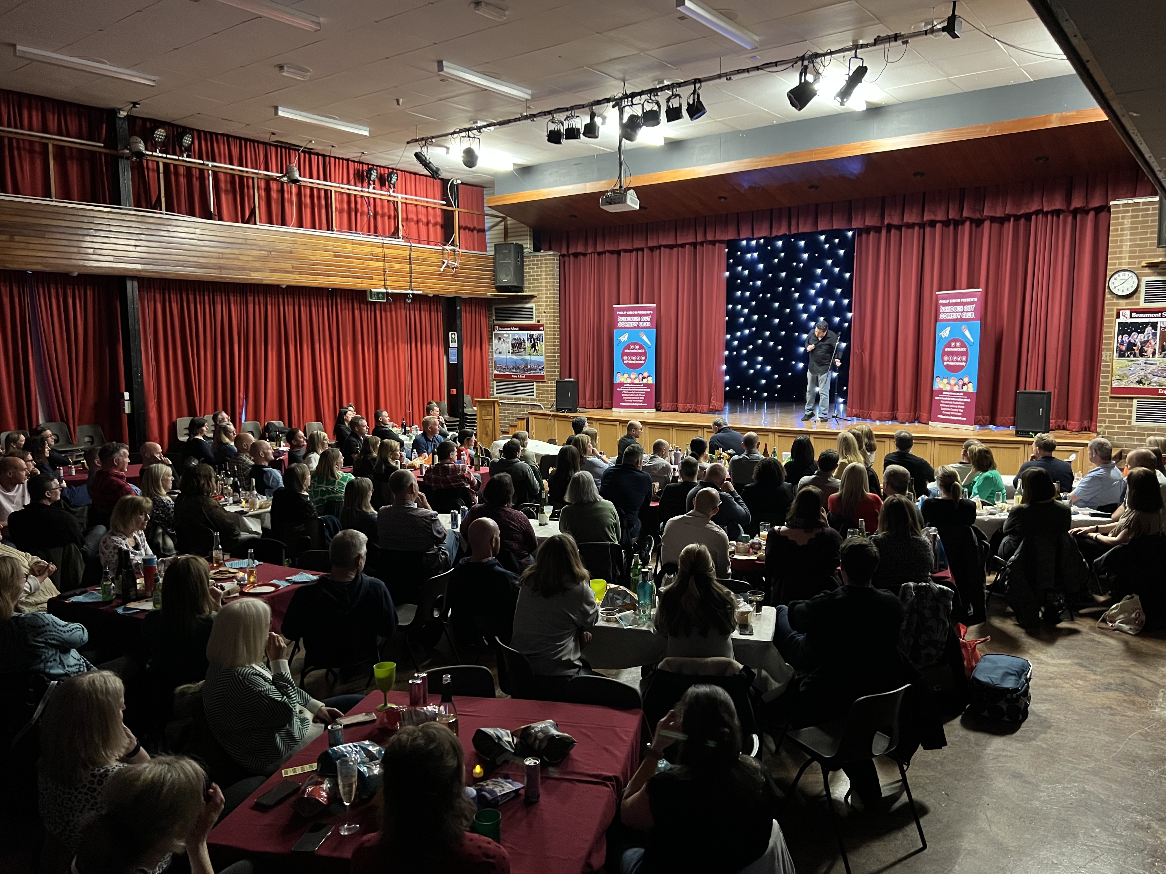 A lively crowd enjoys a comedy show under warm stage lights, with guests seated at tables draped in red cloth, creating a vibrant atmosphere.
