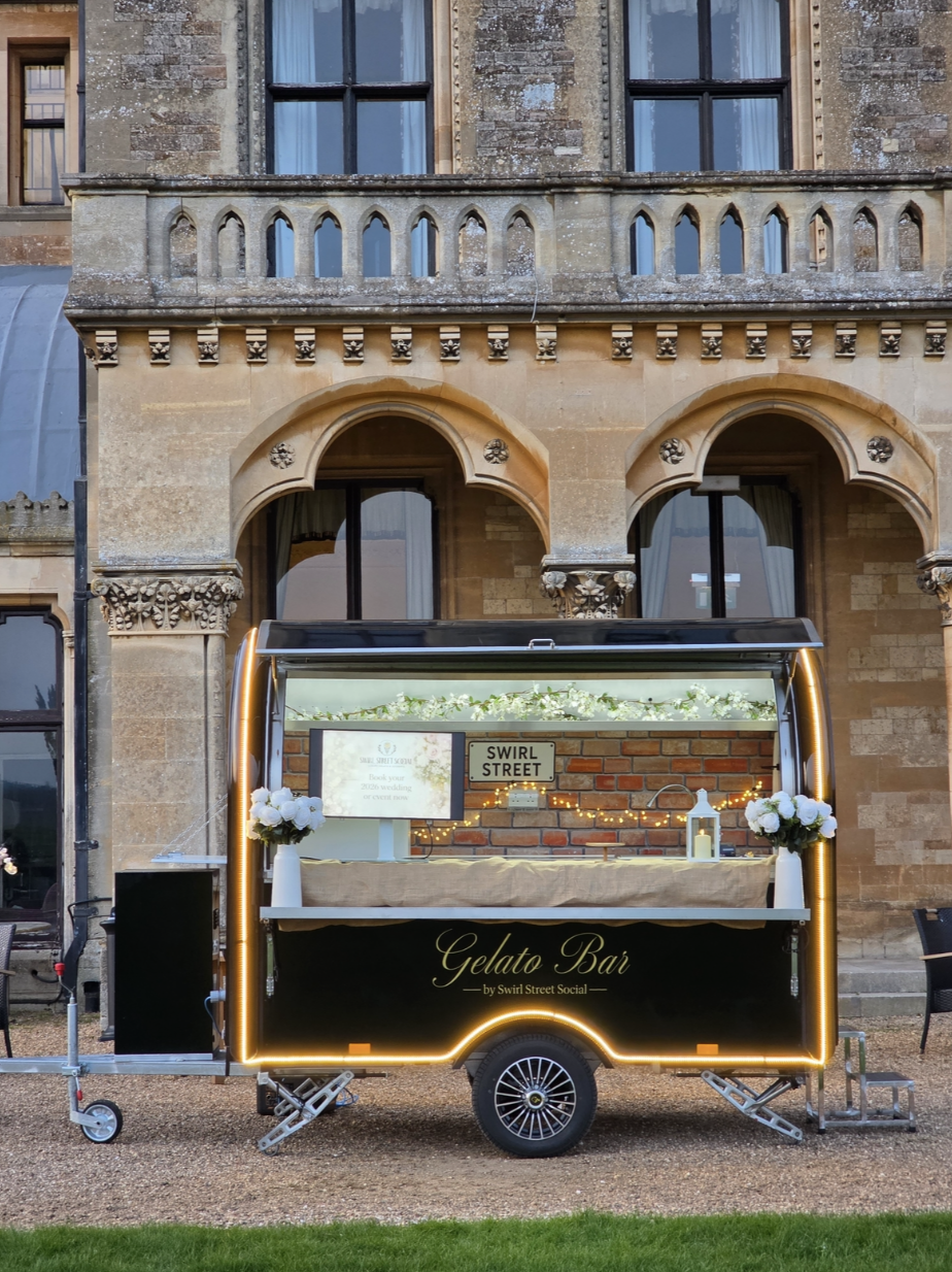 A stylish gelato bar on wheels features elegant decor with white flowers and soft lighting against a stunning stone backdrop, perfect for events.