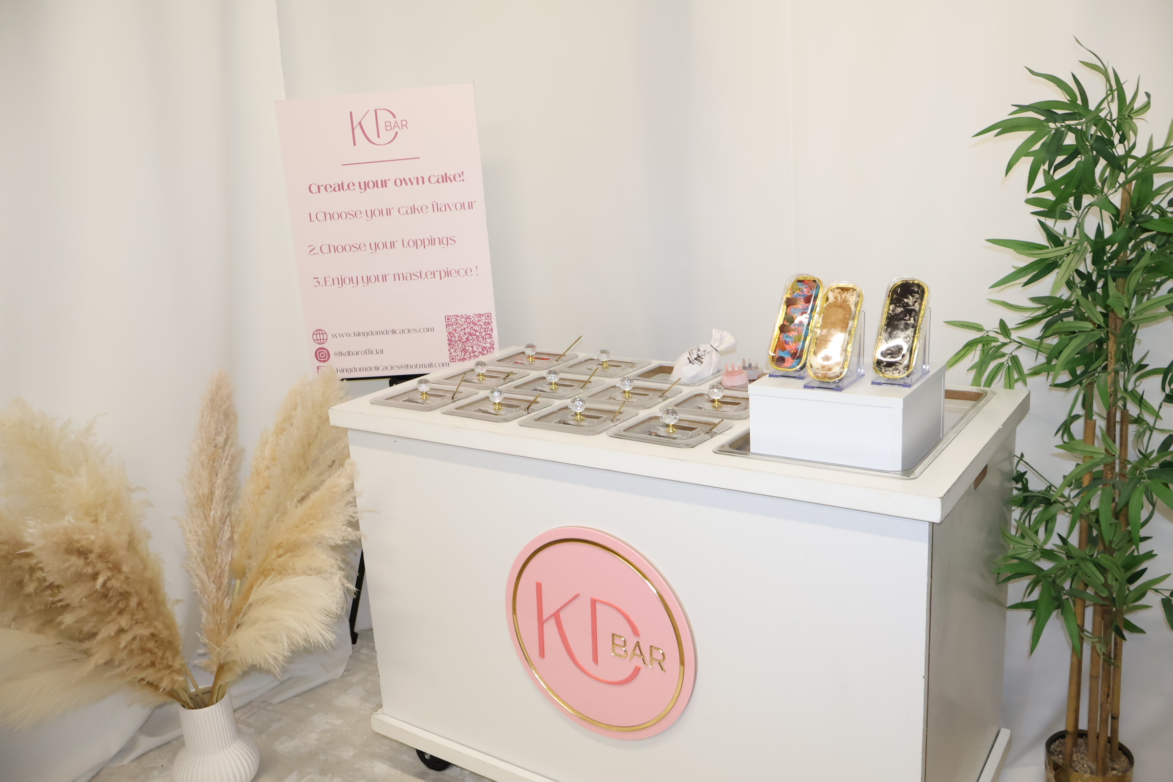 A sleek white cake station features various toppings in clear containers, complemented by a pink sign, creating a cheerful, inviting atmosphere.