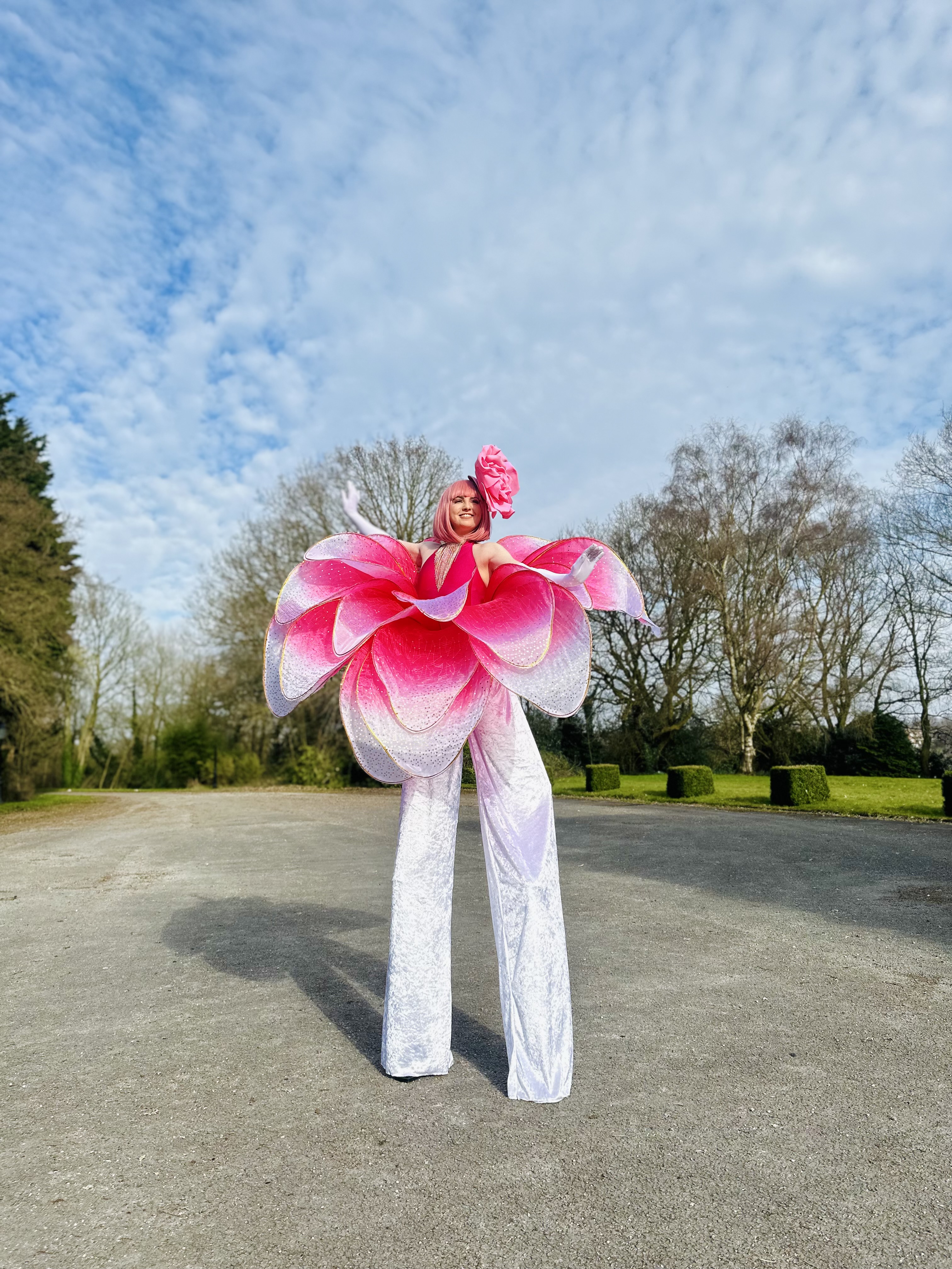 A performer in a vibrant pink flower costume towers on stilts against a bright sky, exuding a whimsical, enchanting festival vibe.