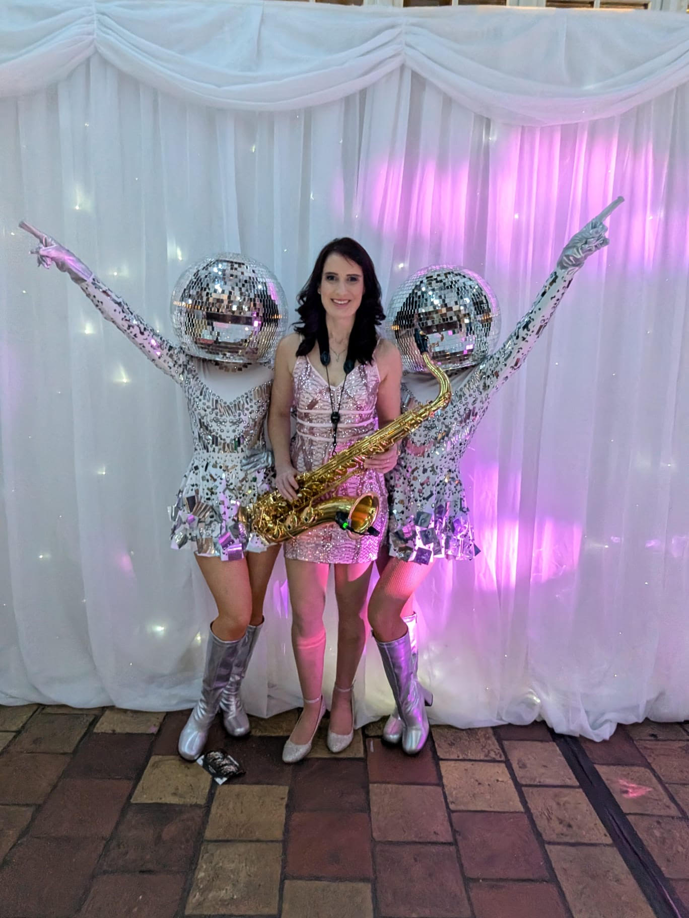 Two dazzling disco-themed performers flanking a saxophonist create a festive atmosphere against a shimmering pink backdrop, perfect for events.