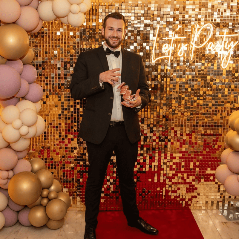 A charming magician in a black tuxedo stands joyfully in front of a sparkling gold backdrop, surrounded by elegant latex balloons.