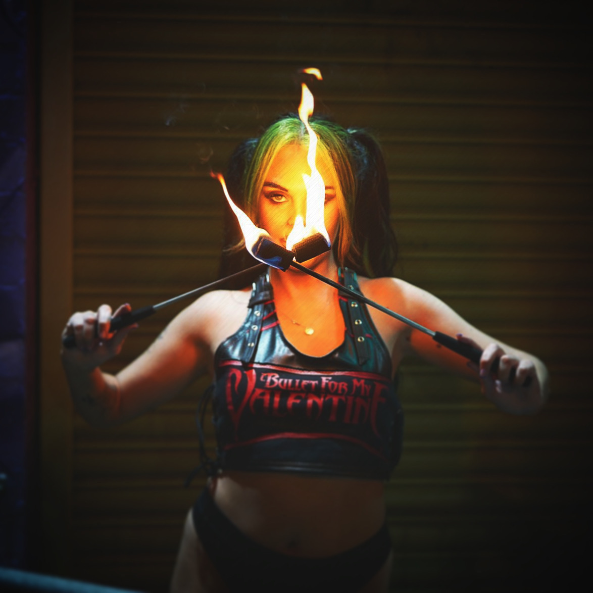 A fire performer with striking green and black hair wields flaming batons, set against a muted backdrop, exuding intense energy and a bold vibe.