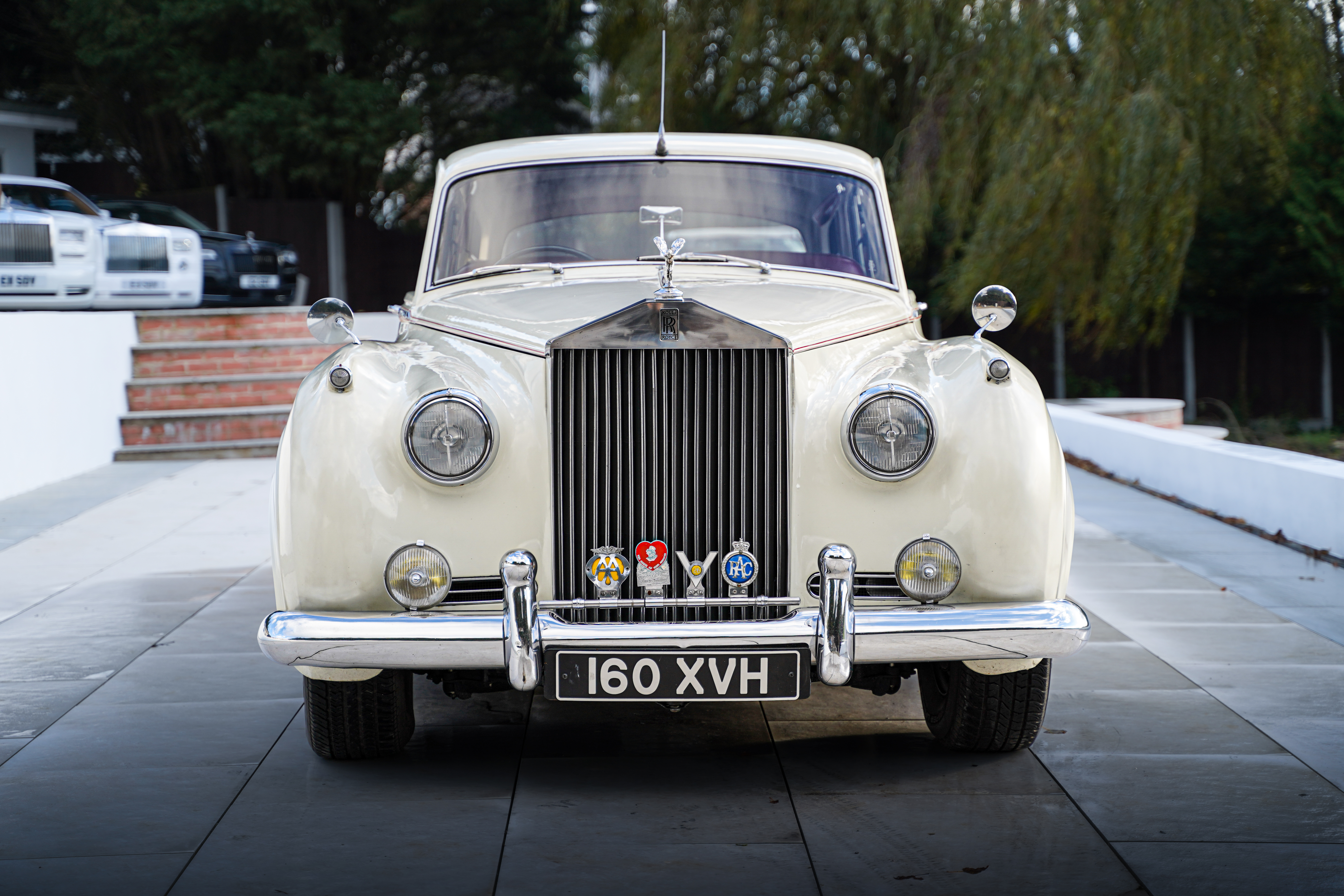 A classic cream Rolls-Royce with polished chrome details sits elegantly on sleek slate tiles, framed by greenery under soft daylight, exuding luxury.