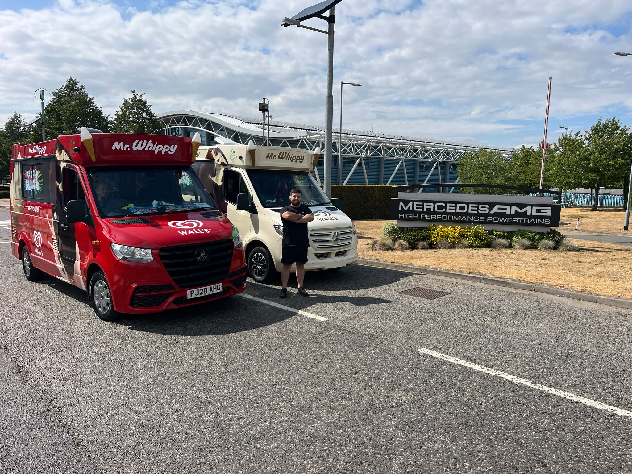 Two Mr Whippy ice cream catering vans, red and cream, parked beside Mercedes AMG signage under bright daylight with a neat roadside setup.