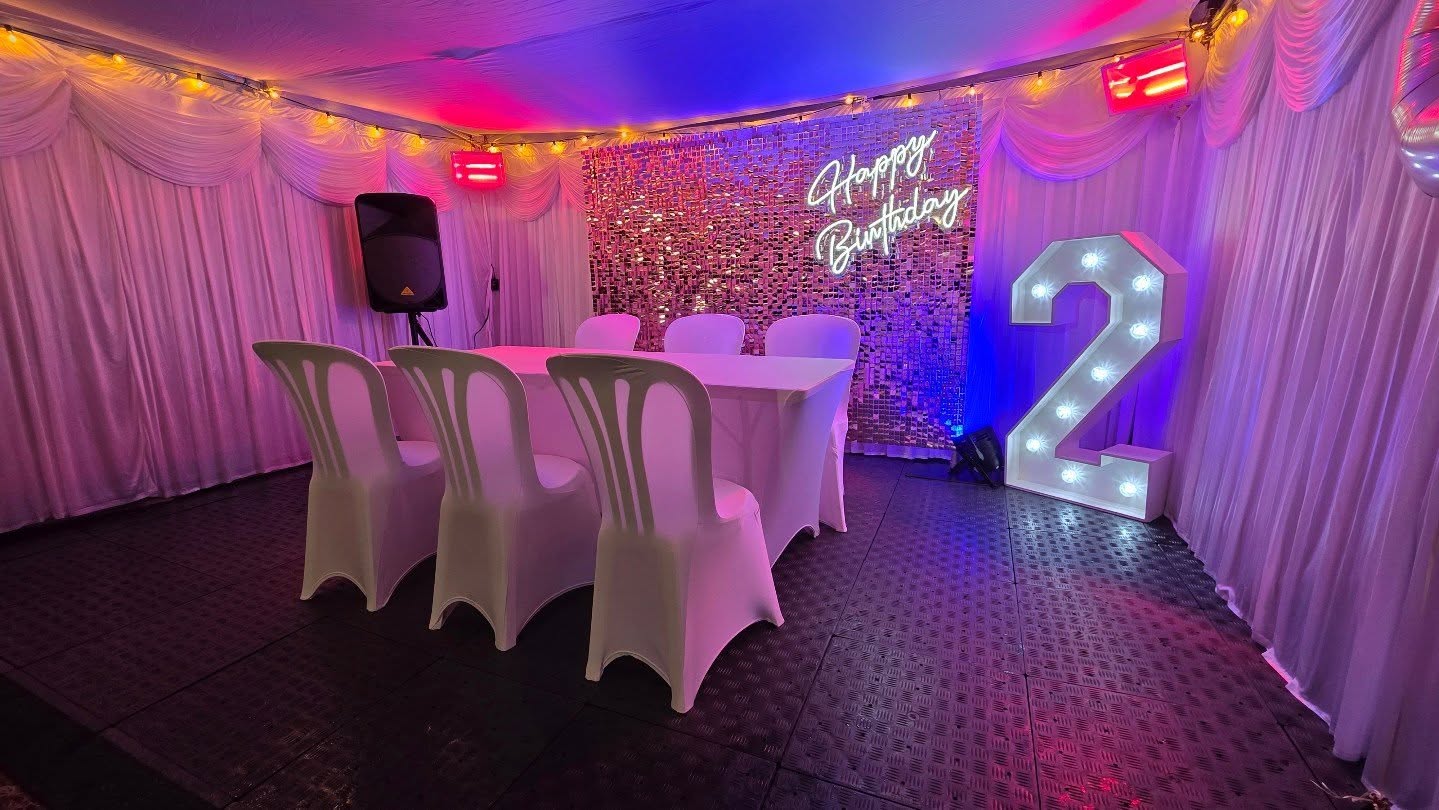 Pink and blue-lit birthday catering setup with white draped table, four covered chairs, sequin backdrop, glowing neon sign, and marquee 2.