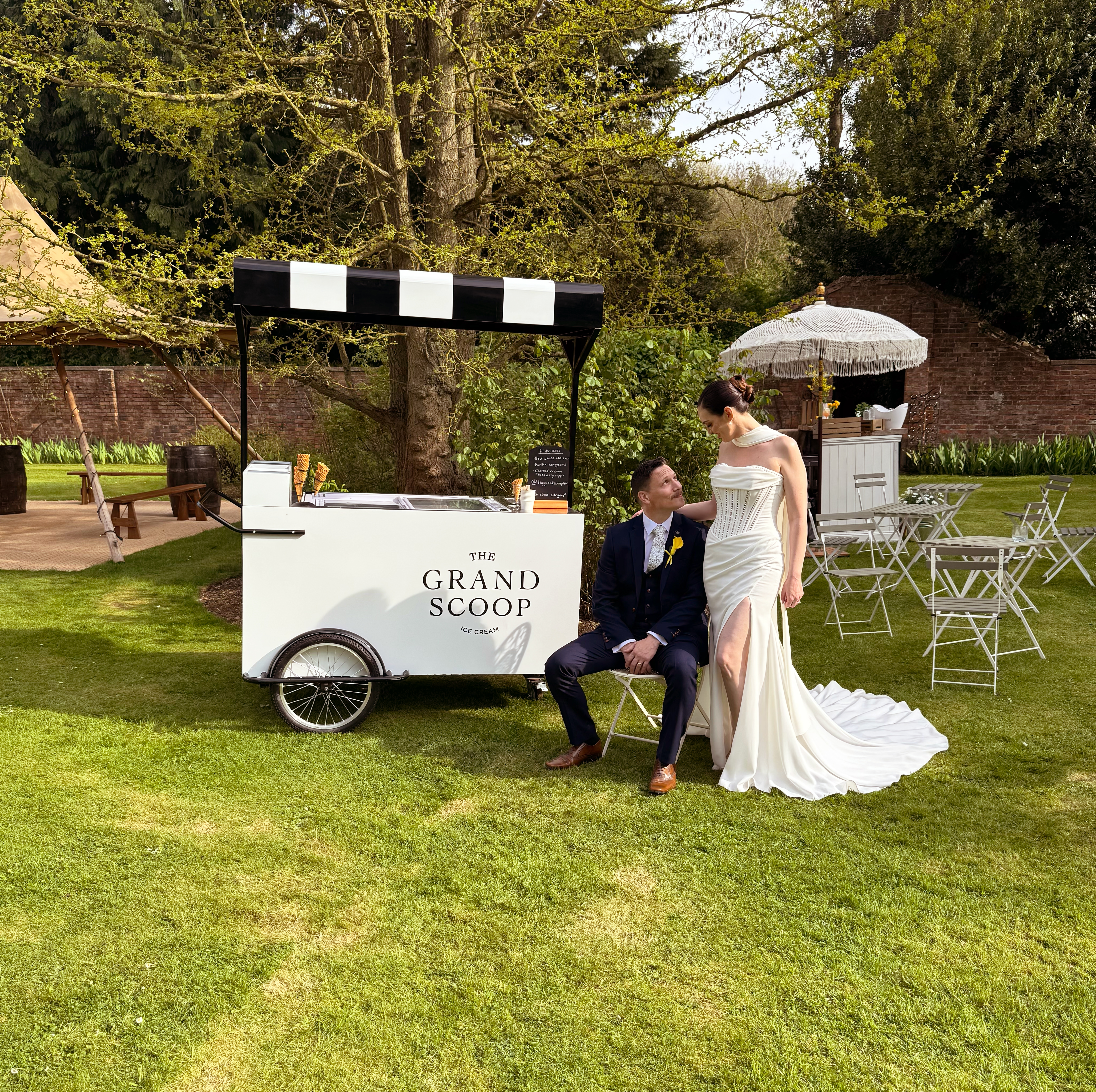 Elegant garden wedding with a vintage ice cream cart, black-and-white canopy, white bistro chairs, and a bride and groom in soft daylight.