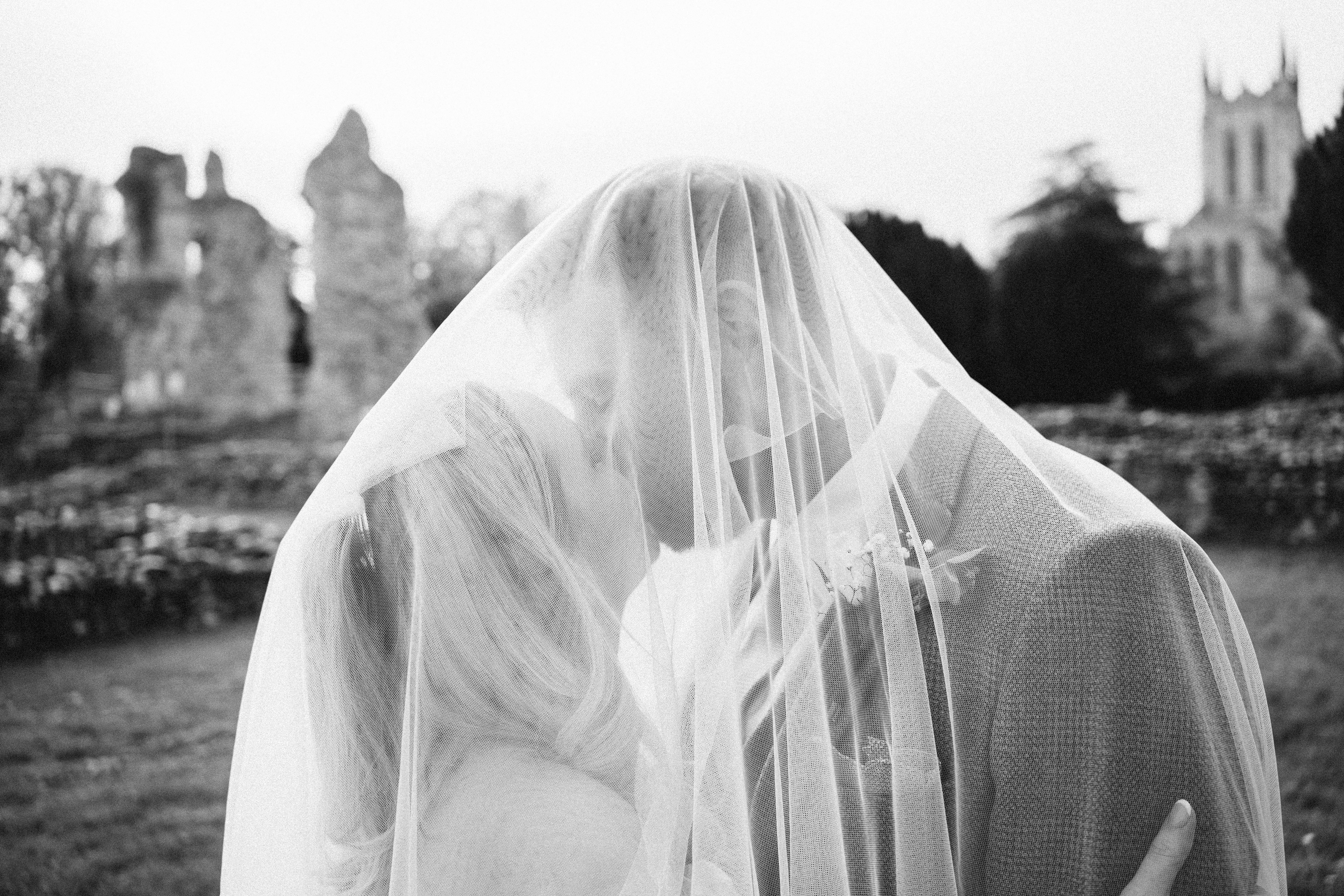 Black-and-white wedding photo of a veiled bride embracing a groom outdoors, with soft daylight and a blurred historic church backdrop.